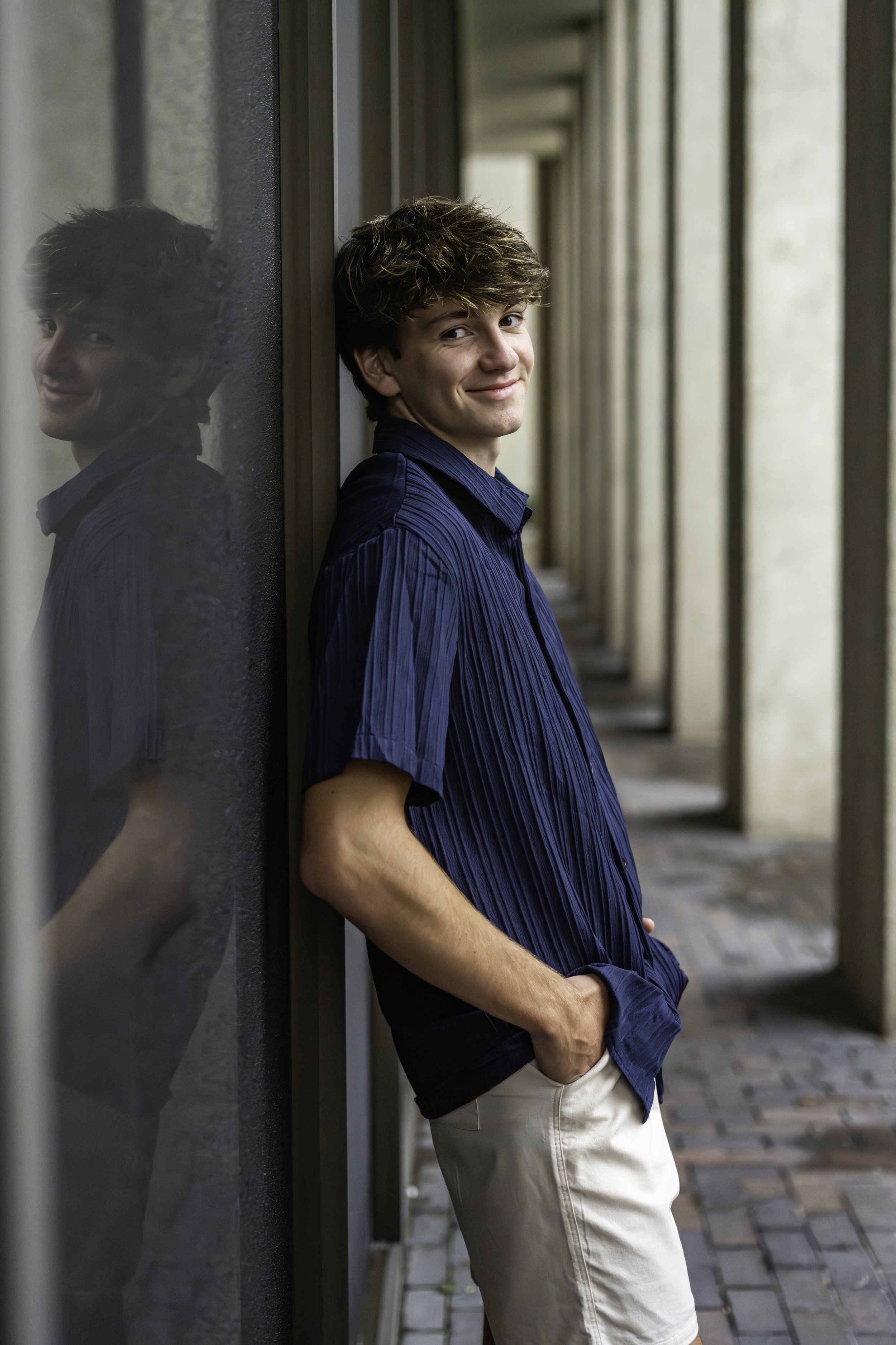 A young man with brown, curly hair smiling at the camera while leaning against a glass wall, with his hands in his khaki shorts' pockets, standing outdoors on a brick-paved walkway.