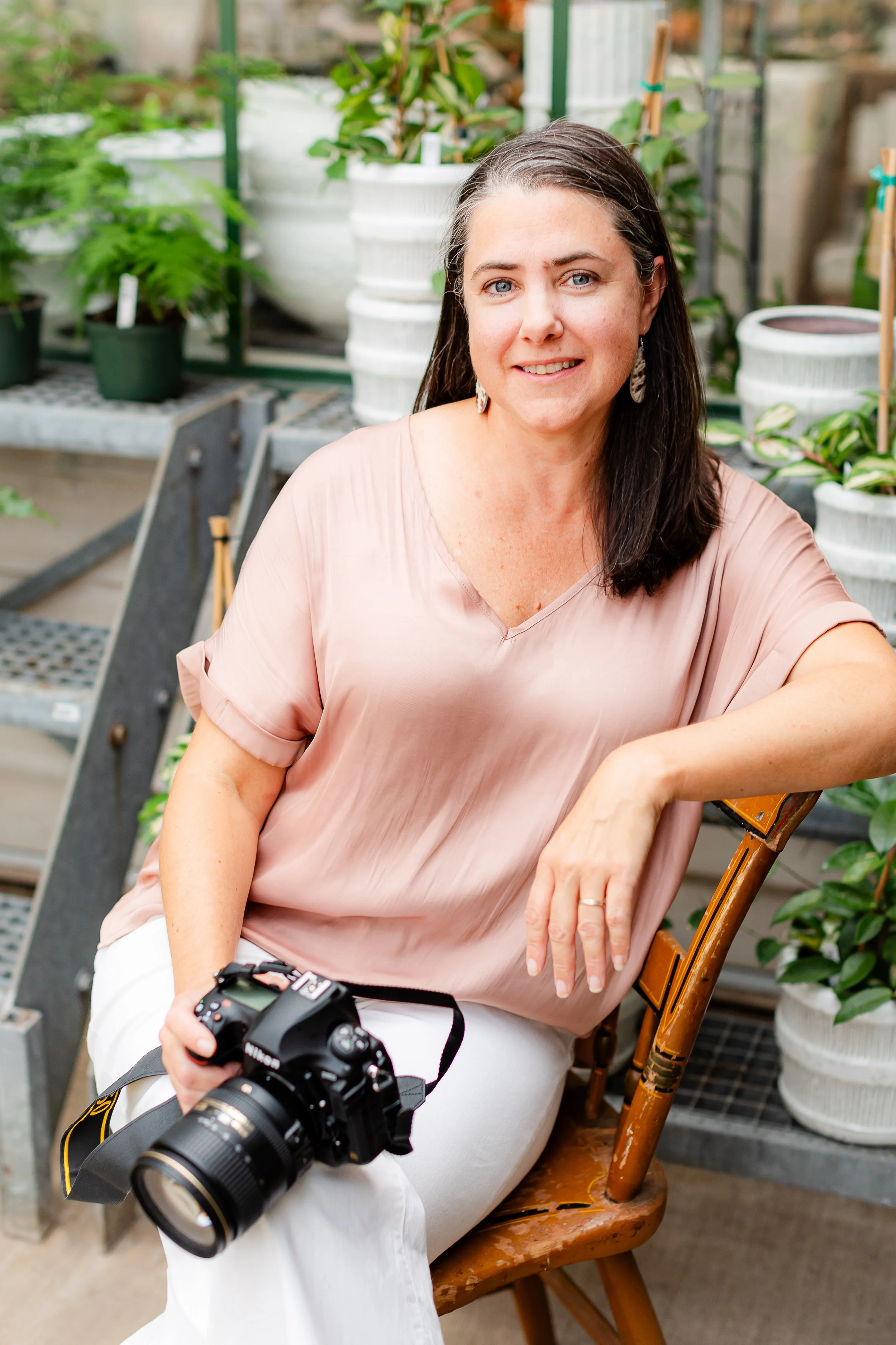 Kristie Allen with dark hair, wearing a light pink blouse and white pants, sitting on a wooden chair inside a green plant nursery. She is holding a camera in her lap, smiling at the camera.