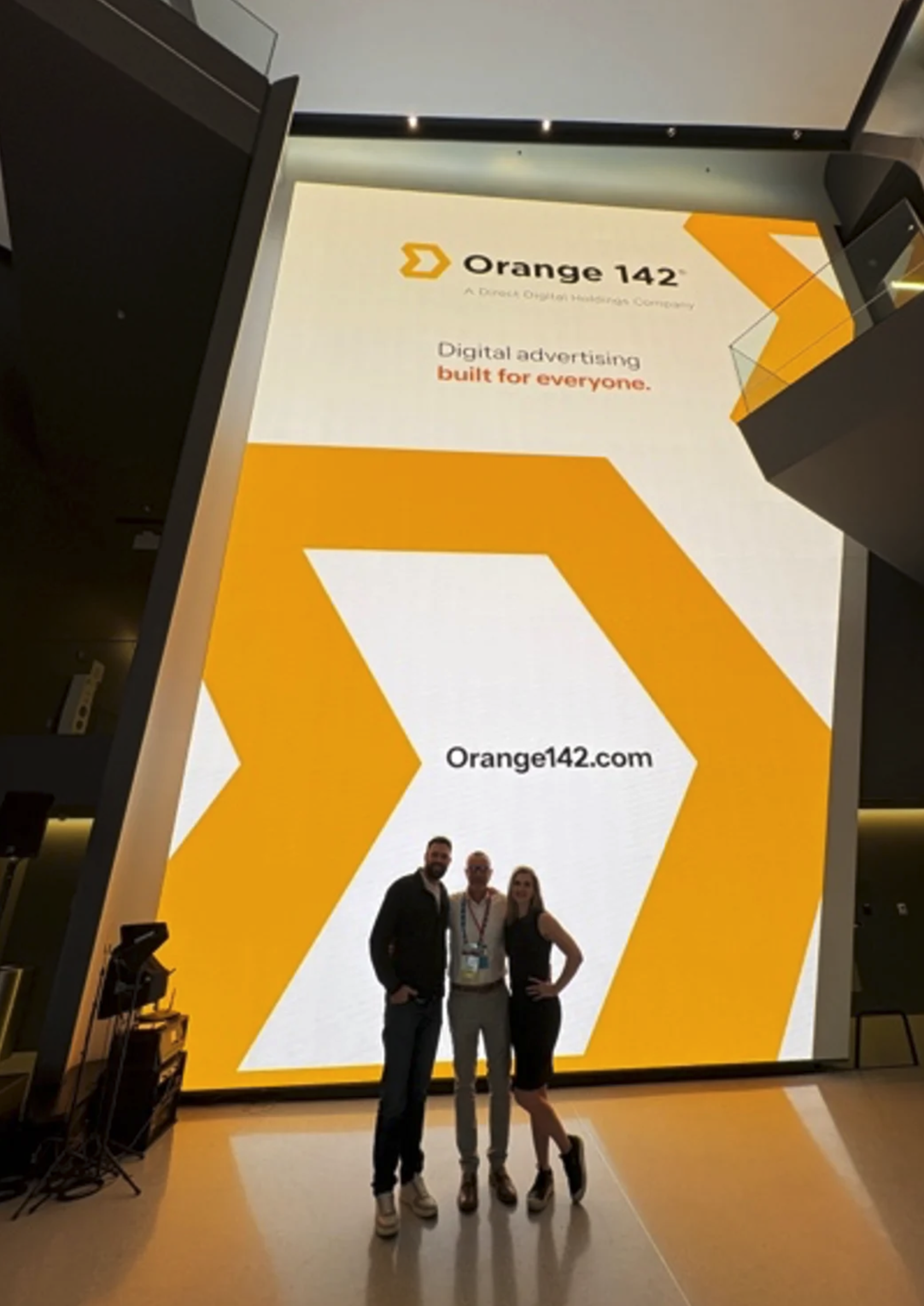 Three people standing in front of a large digital screen displaying advertising for Orange 142, a digital advertising company. The screen features branding in orange and white, with a website URL 'Orange142.com' and the tagline 'Digital advertising b