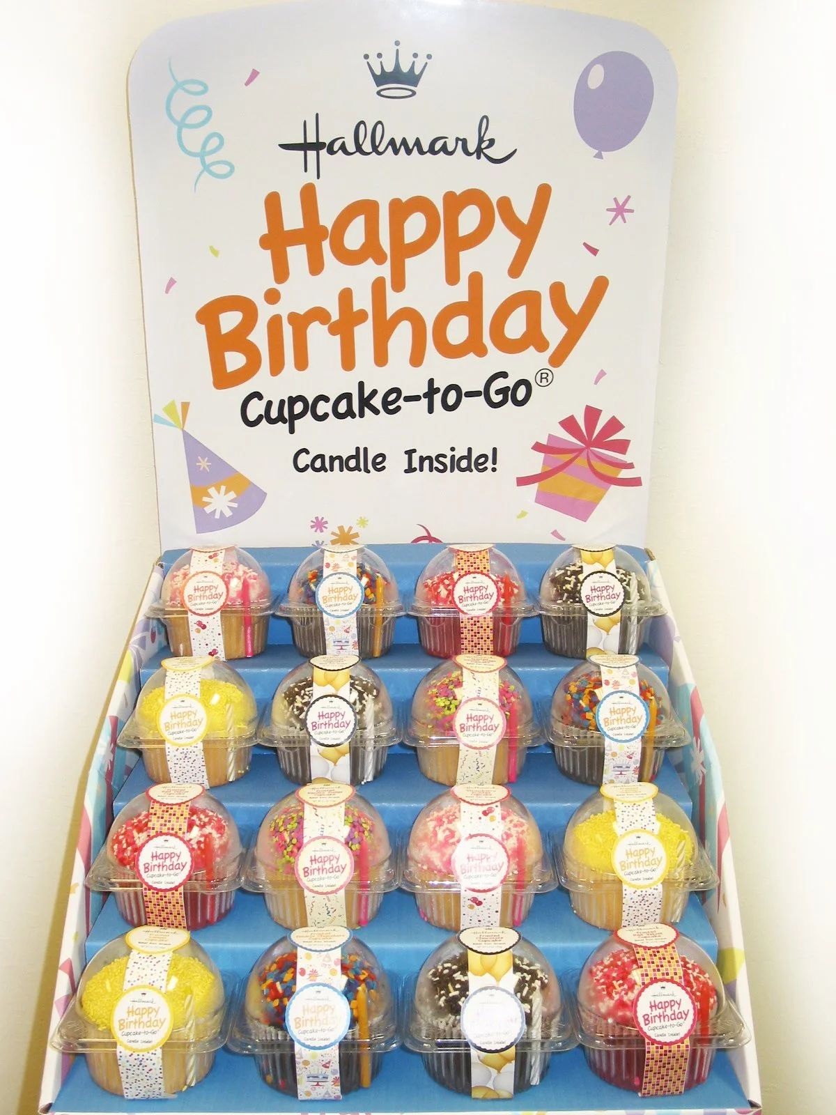 Display box of small cupcakes with colorful frosting and sprinkles, each with a 'Happy Birthday' label, inside a retail setting.