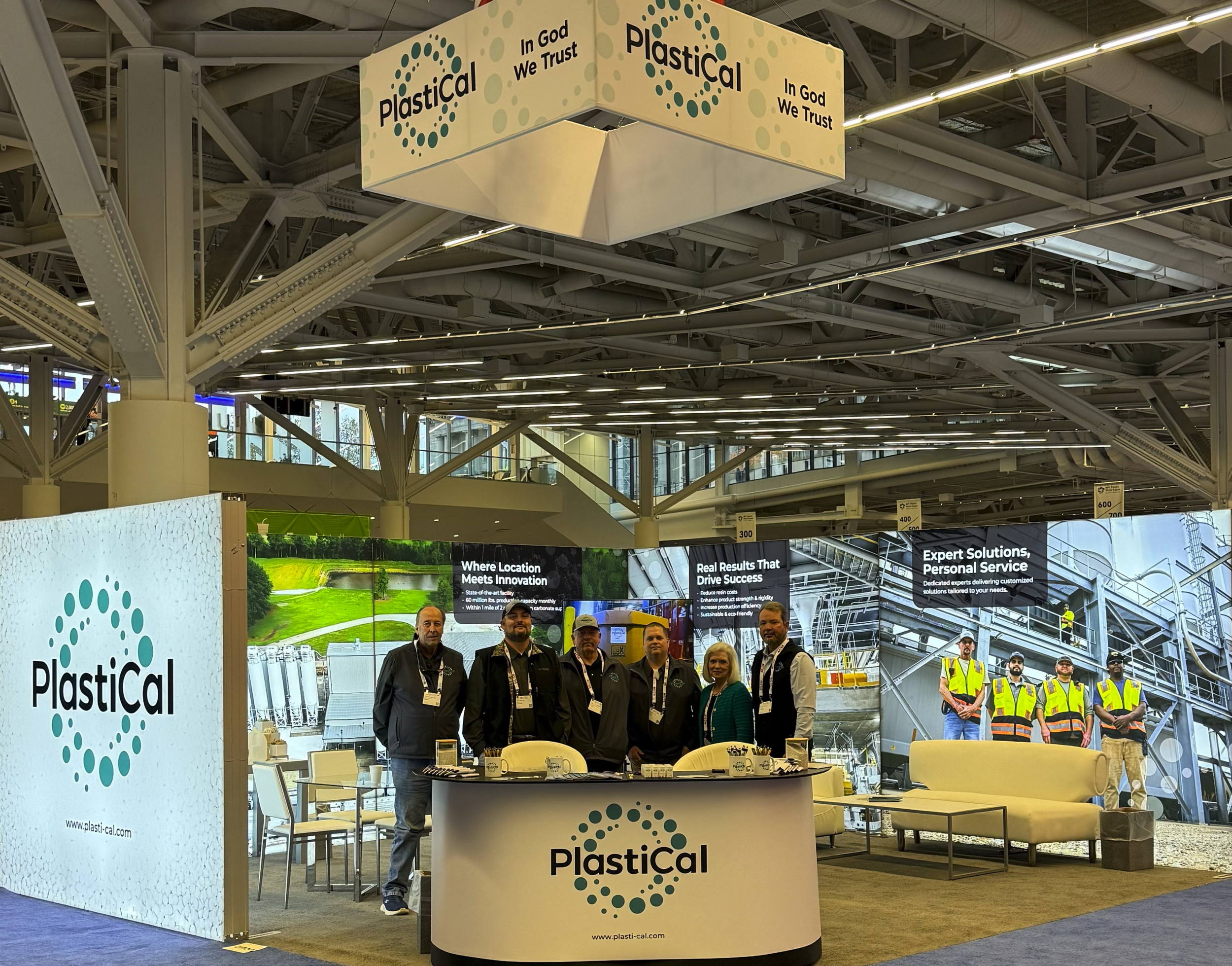 Trade show booth for a company named PlasticCal, with a group of six people standing behind a curved desk, large digital screens in the background, and promotional signs about innovation, real results, expert solutions, personal service, and safety.
