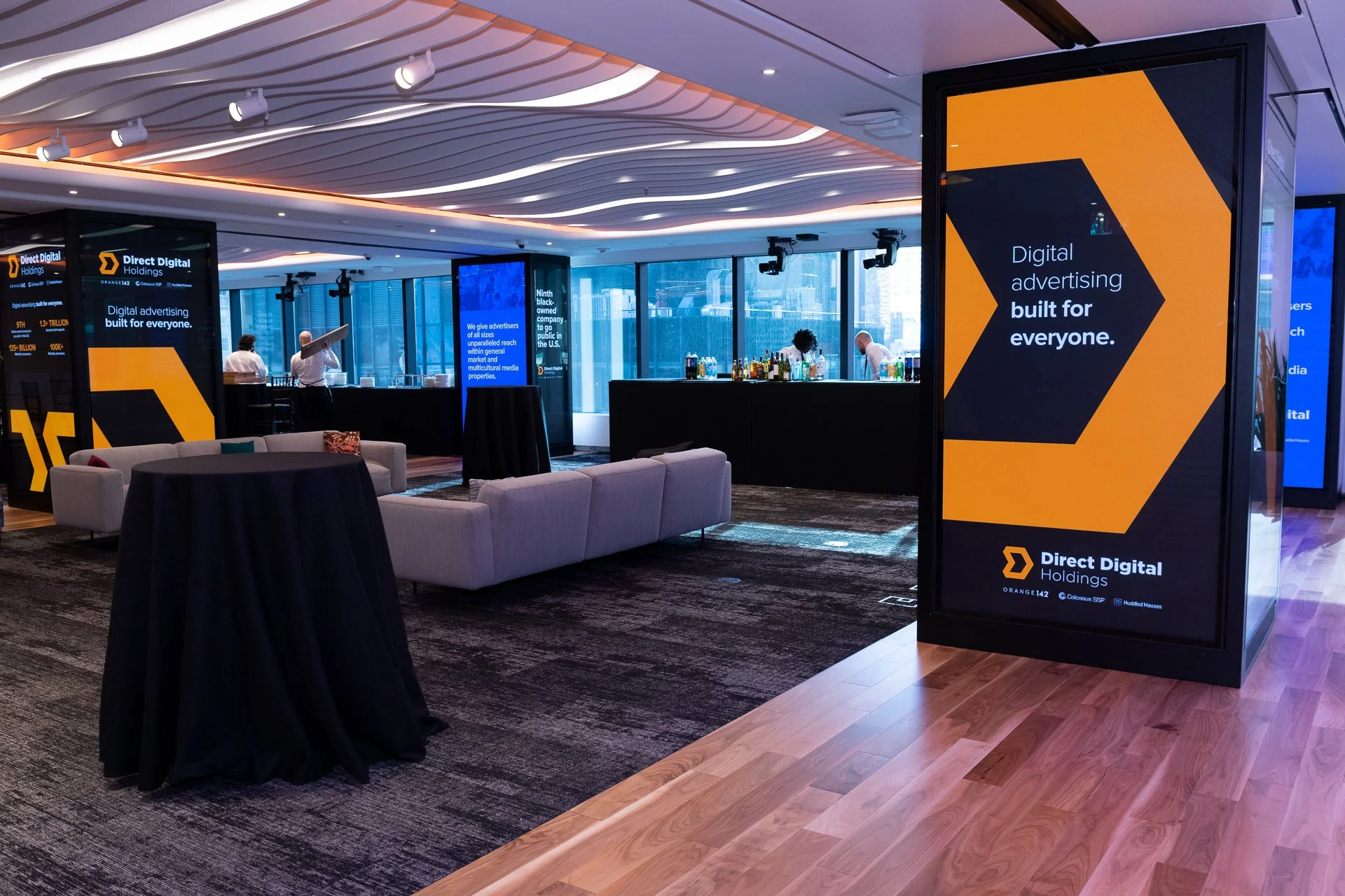 Interior of a modern office lounge with digital advertising displays, a bar area with bottles, some couches, and a table, with large windows in the background.