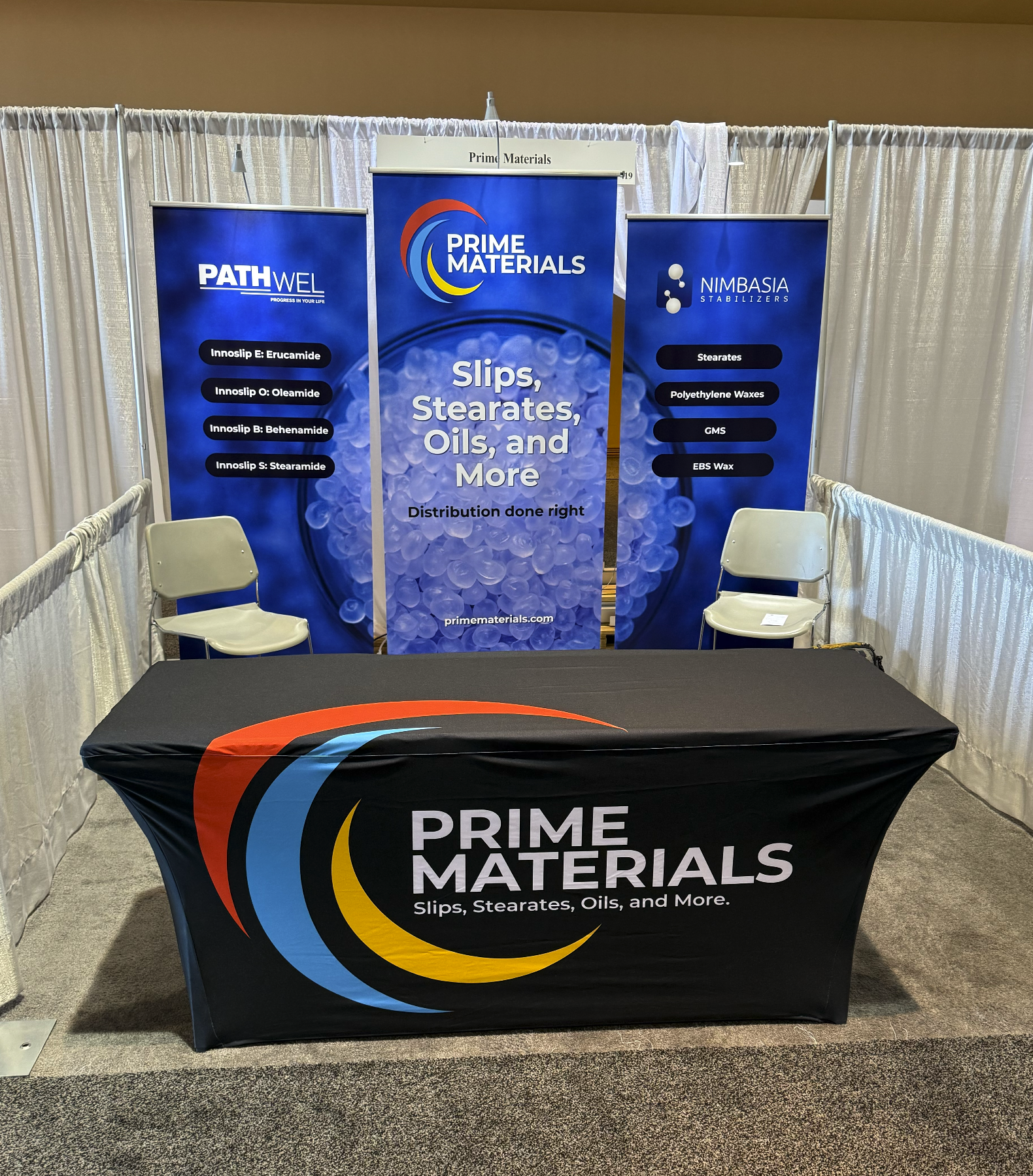 A trade show booth for Prime Materials featuring a table with a black tablecloth and the company logo, and three vertical banners advertising slips, stearates, oils, and more, with two chairs in front of the booth.