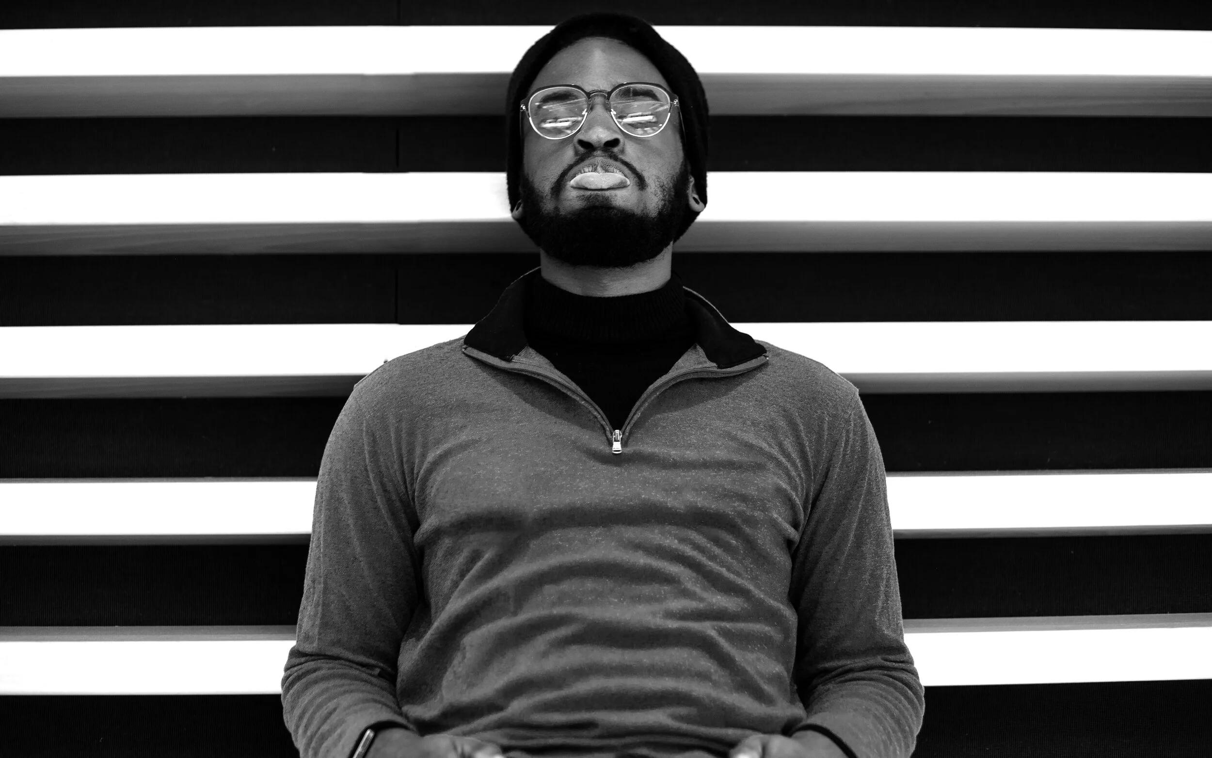 Black and white photo of a person wearing glasses and a beanie, standing in front of horizontal light strips.