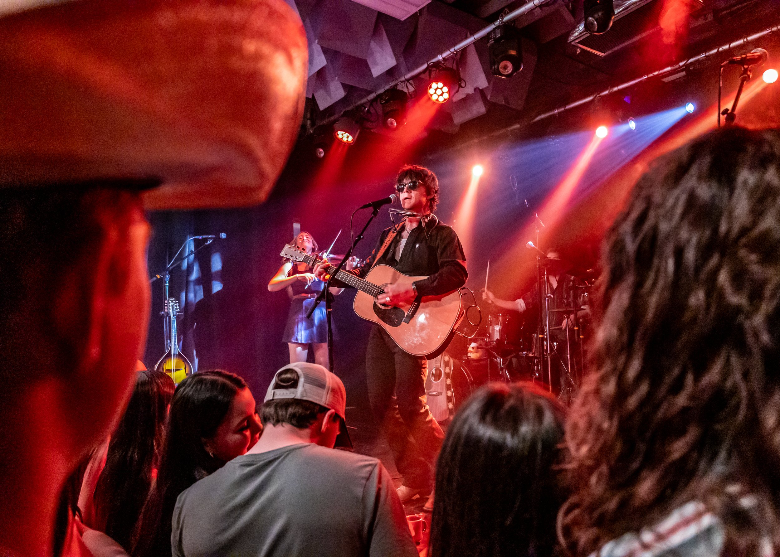 A musician performing on stage with a guitar, wearing sunglasses and surrounded by colorful stage lights, with a crowd in the foreground.