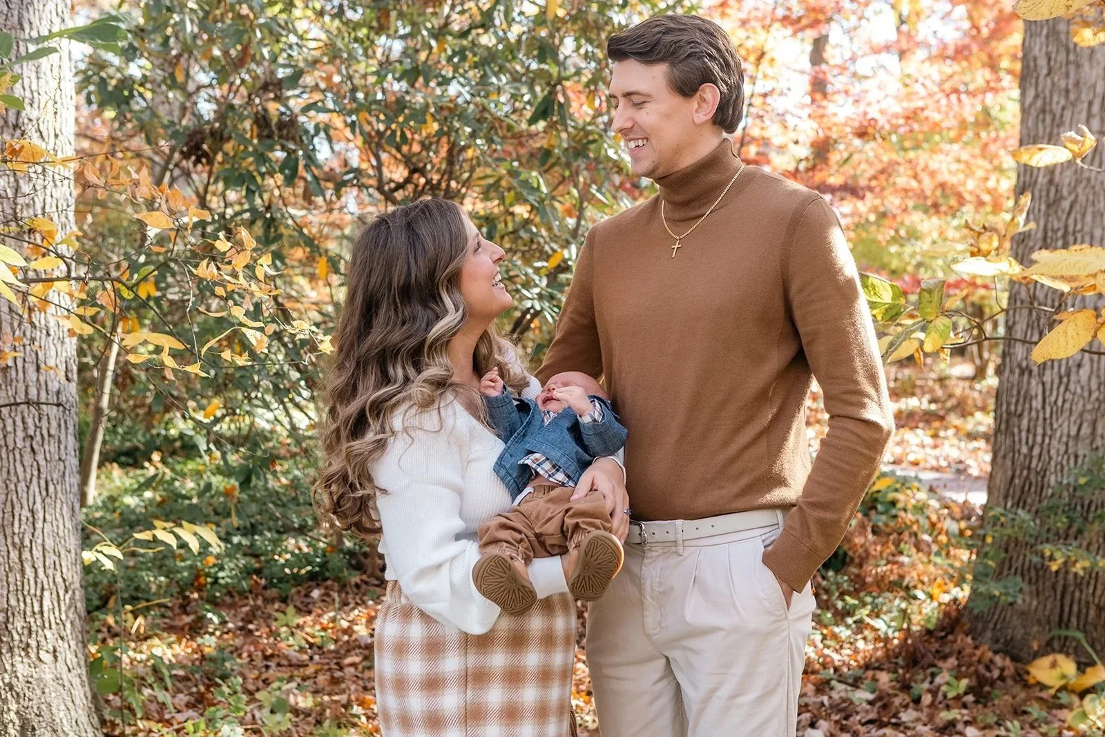 A smiling woman holding a baby and looking up at a man. The man is smiling back at her in a forest with autumn leaves.