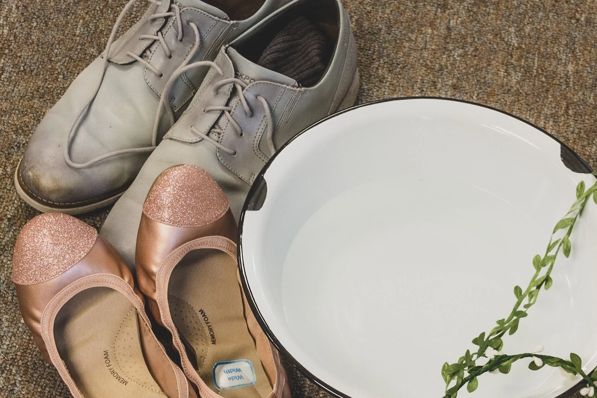 A grey pair of shoes and a pink pair of glittery slippers next to a bowl with a green branch
