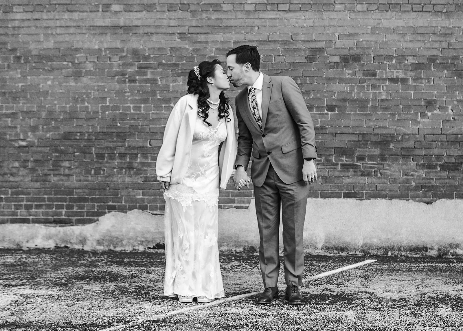 A black-and-white photo of a couple holding hands, softly kissing, standing in front of a brick wall on an asphalt surface, dressed in wedding attire.