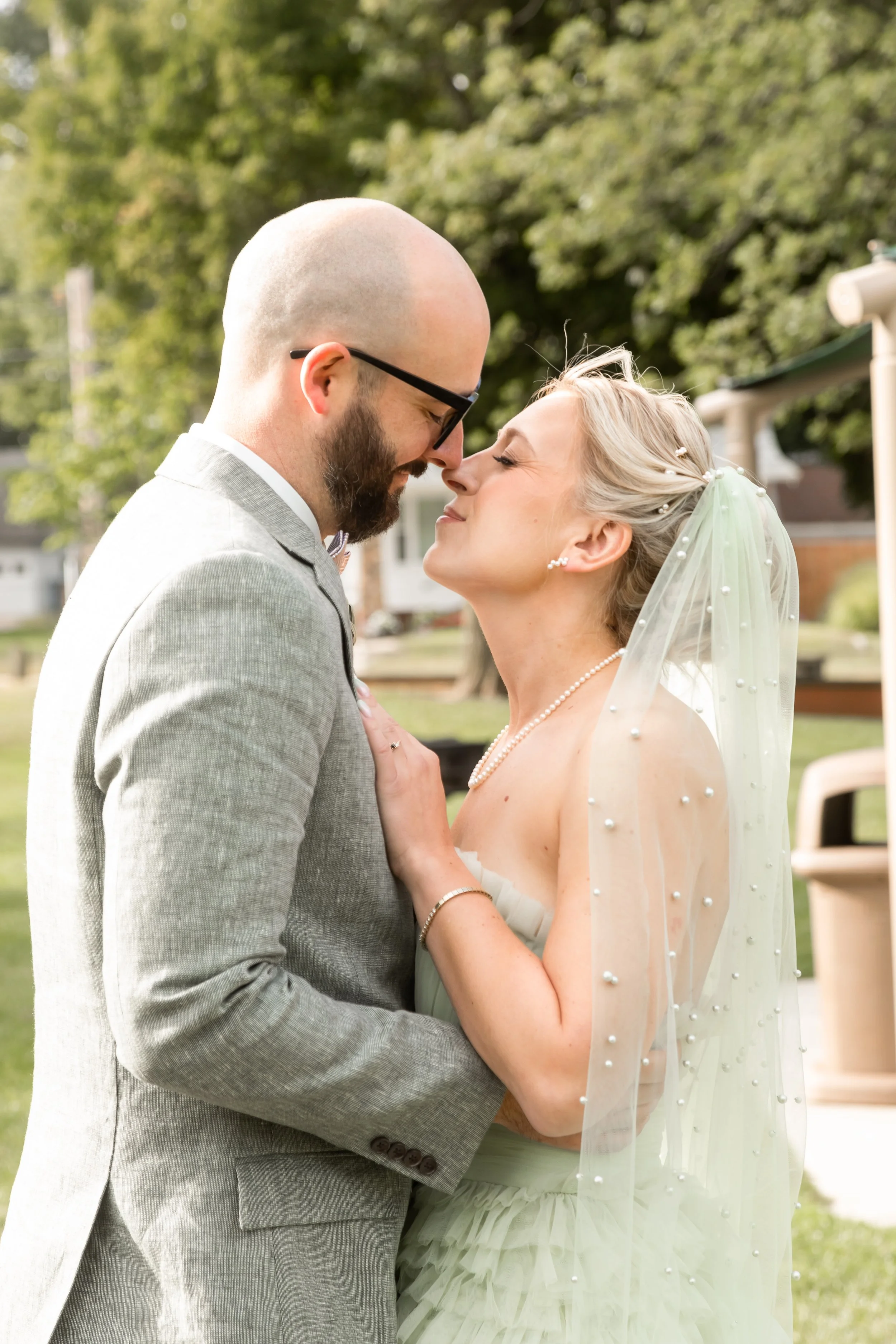 A loving couple shares a close moment during their wedding outdoors, with a background of trees and houses.