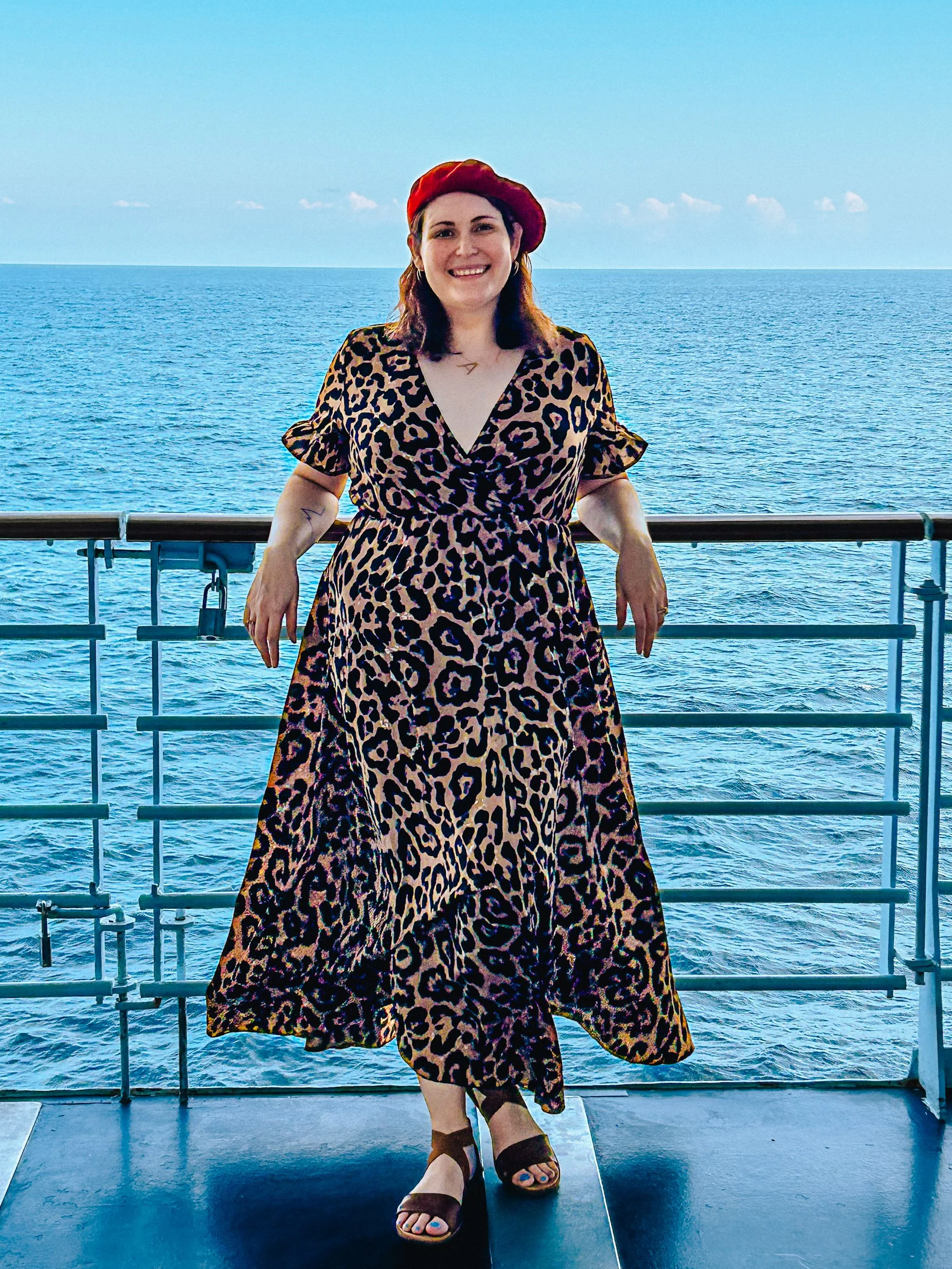 A woman wearing a leopard print dress and red beret standing on a ship deck with the ocean in the background.