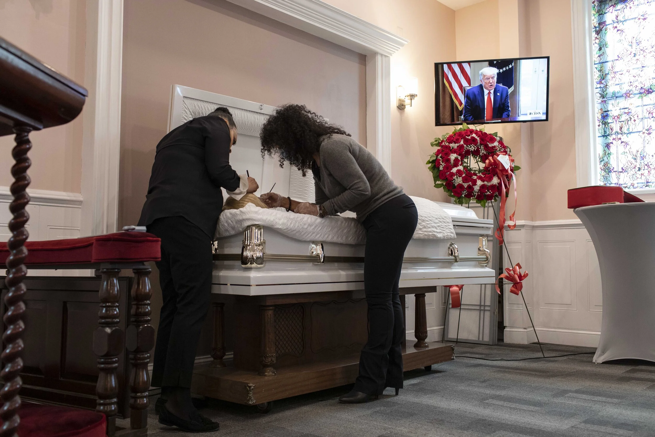  Jenny Adames, left, applies make up to her aunt, a victim of COVID-19, before her viewing service as a screen plays a press conference held by U.S. President Donald Trump.  