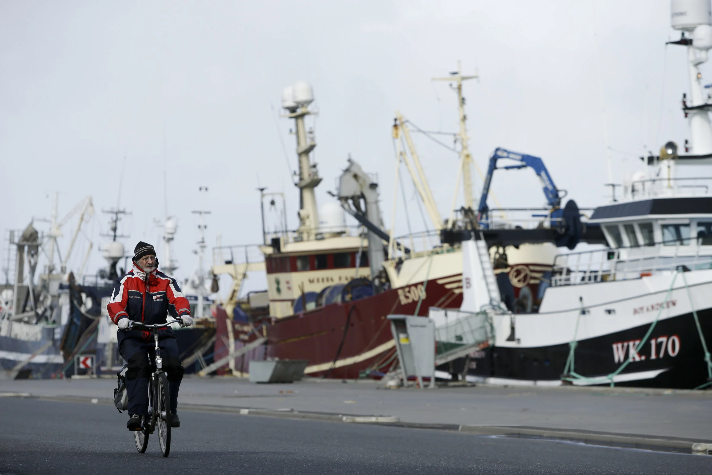  A person rides a bicycle by docked fishing ships while wearing coveralls. Given the intermittent weather, such clothing is seemingly omnipresent in the area for men women and children. Some refer to it as the West Coast Tuxedo.  