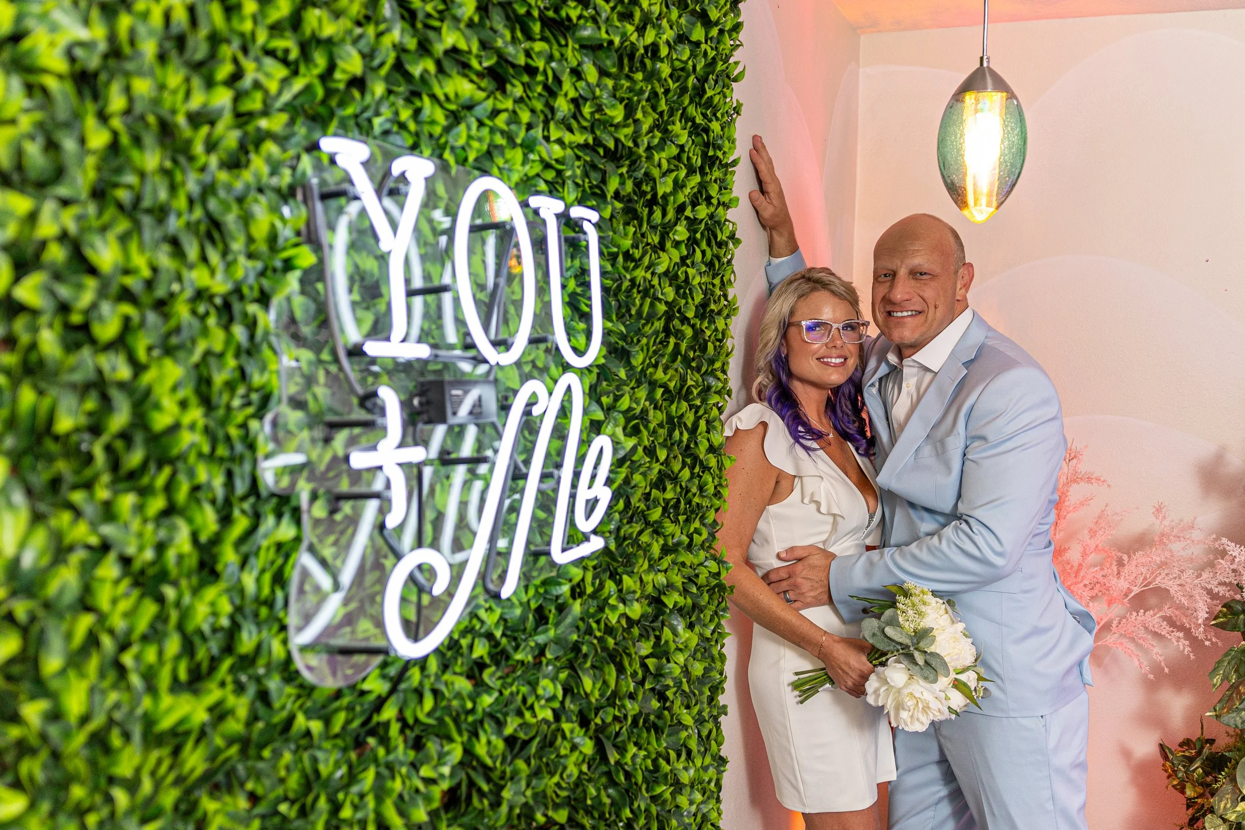 Loving couple hugging by a green way with a neon sign that says you + me