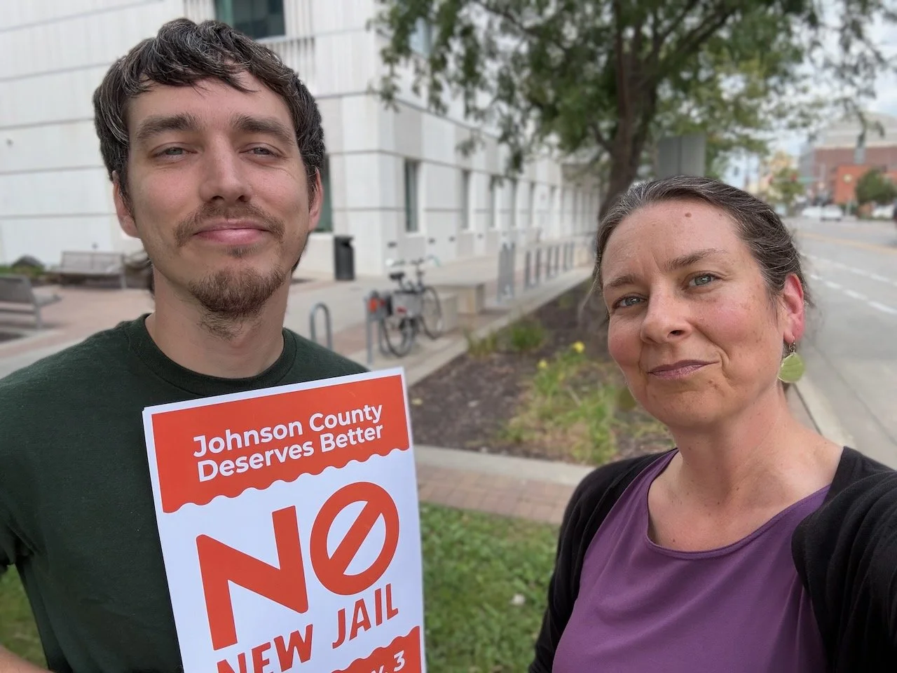 Oliver Weilein and Laura Bergus pose for a selfie on S. Clinton Street. Oliver is holding a "No New Jail" sign from the Johnson County Deserves Better campaign.
