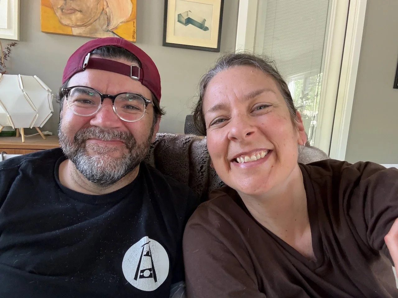 Nick and Laura Bergus smile for a selfie. They're seated; Nick wears a backwards baseball cap and glasses. Laura's hair is pulled back and she's in a brown v-neck t-shirt.
