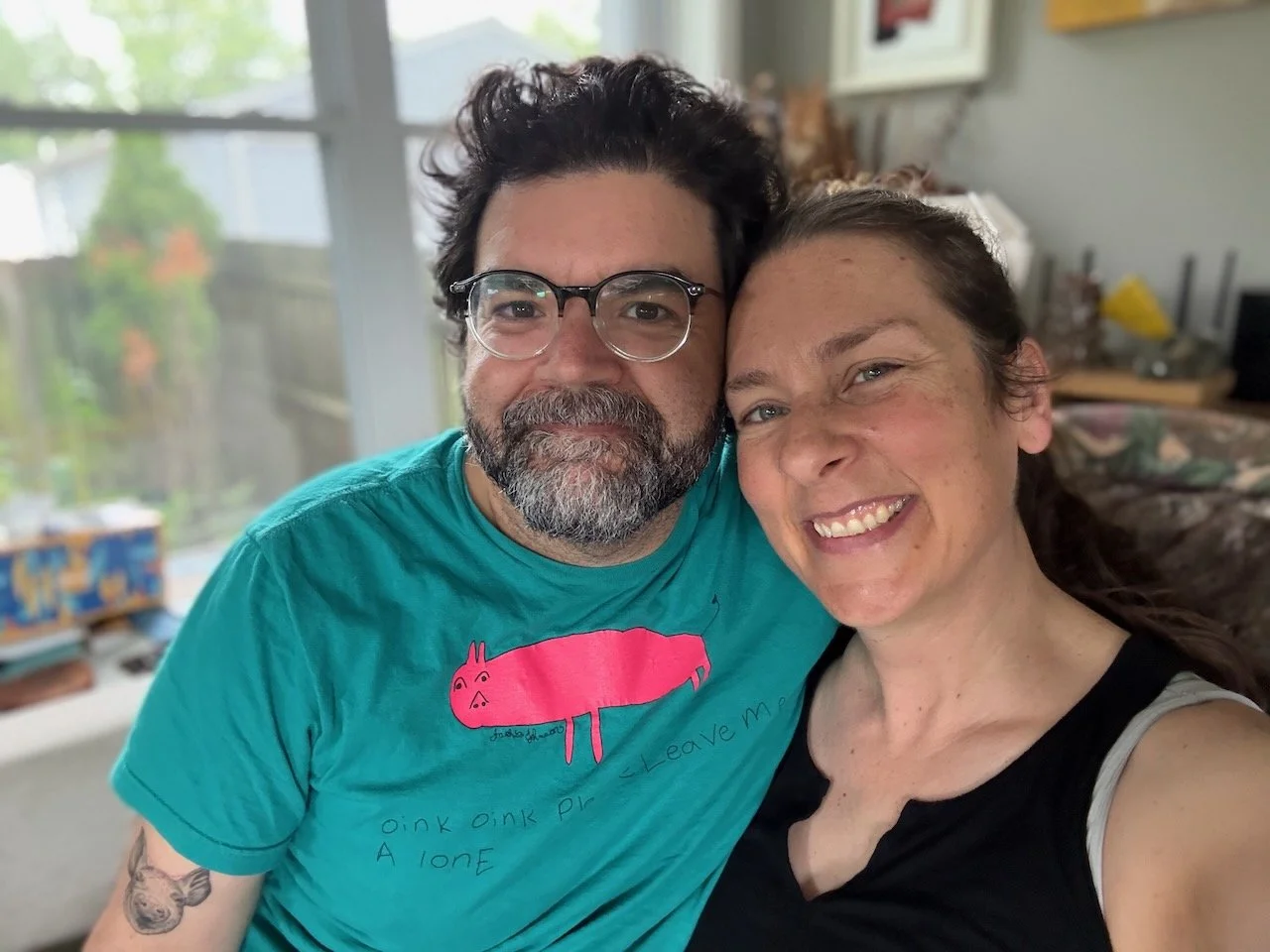 Nick Bergus and Laura Bergus smile at the camera. They are seated on a couch.