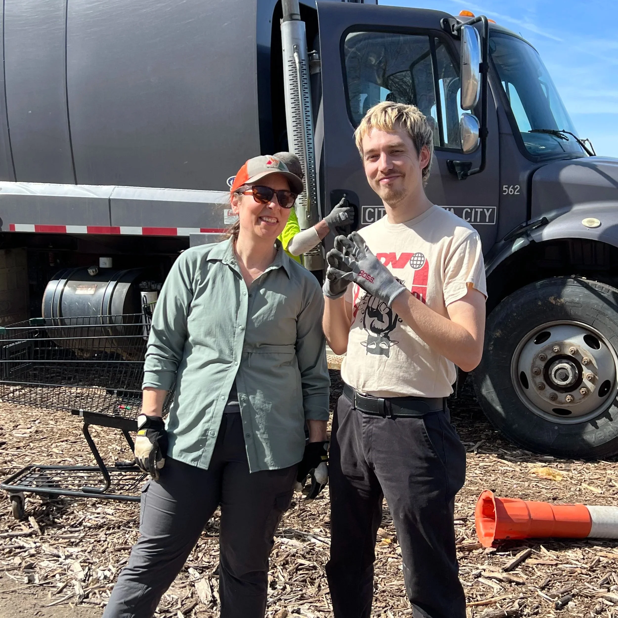 Oliver Weilein and Laura Bergus pose standing in front of a City of Iowa City garbage truck, near a shopping cart and traffic cone.