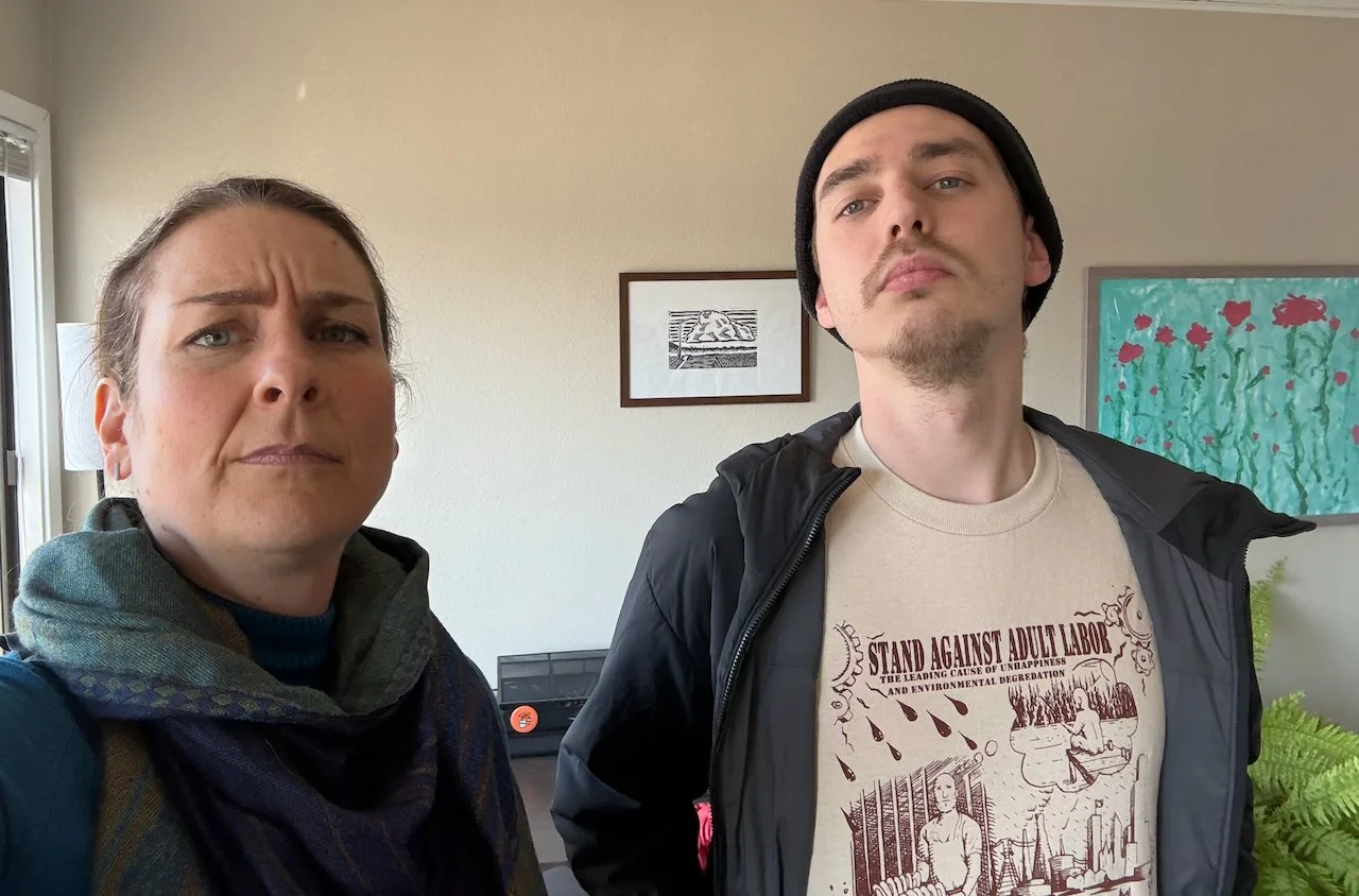 Laura Bergus scowls while Oliver Weilein shows off a t-shirt with the words "Stand Against Adult Labor" across the chest. Art by Casey Hayes and Nick Bergus in the background.