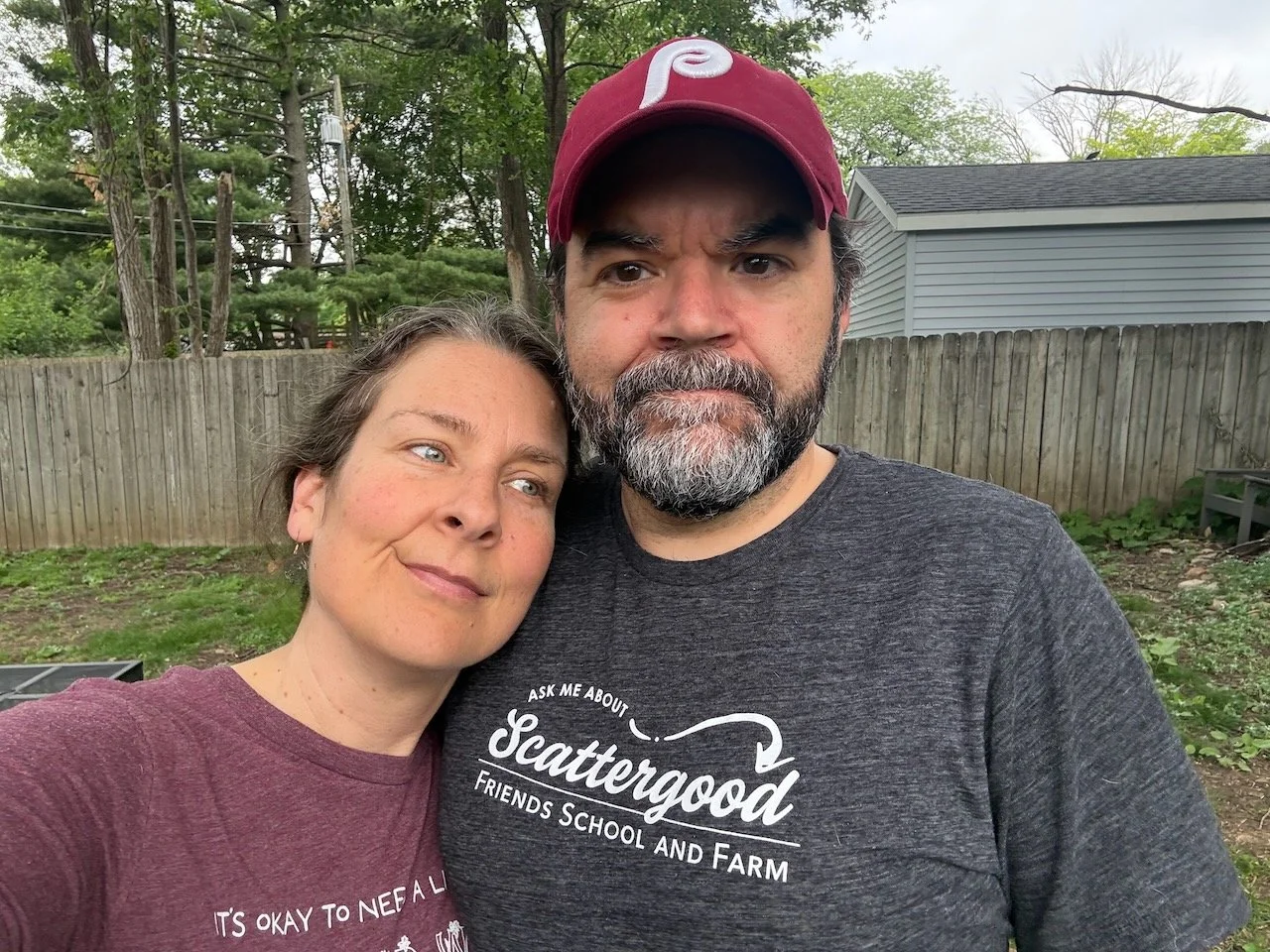 Laura Bergus and Nick Bergus pose for a selfie in front of a wooden privacy fence. A scruffy, weedy yard and neighborhood trees are in the background.