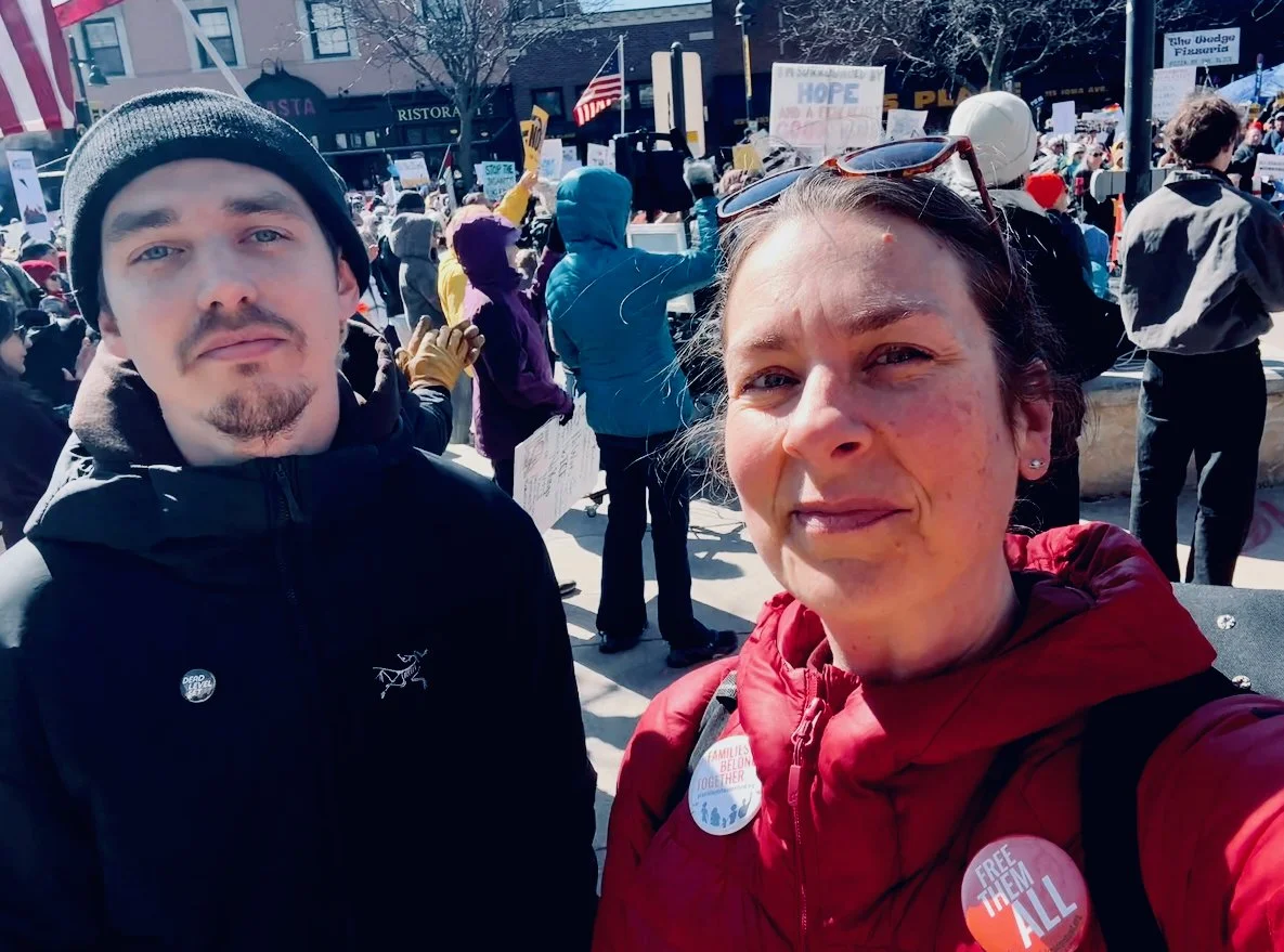 A white man in a black jacket and stocking cap pose with a white woman in front of a crowd holding up signs protesting fascism.
