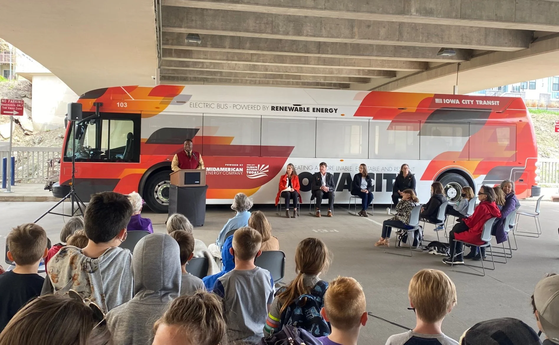 One of Iowa City Transit's electric buses on display during an event with Mayor Bruce Teague at a podium and transit officials and a crowd of the public in front of the bus.