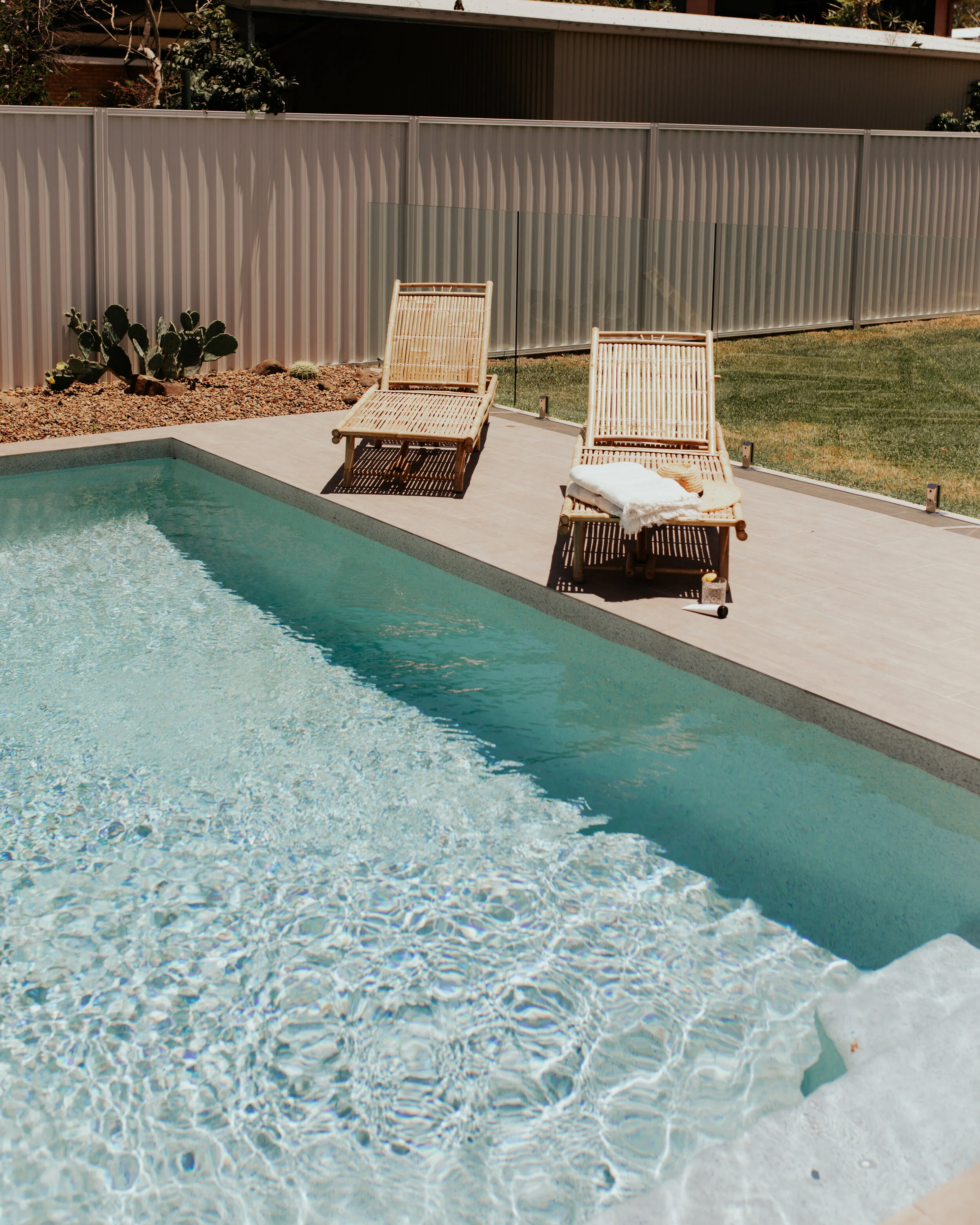 Two bamboo lounge chairs with towels and a small cup on a poolside patio, overlooking a clear backyard swimming pool at a holiday rental