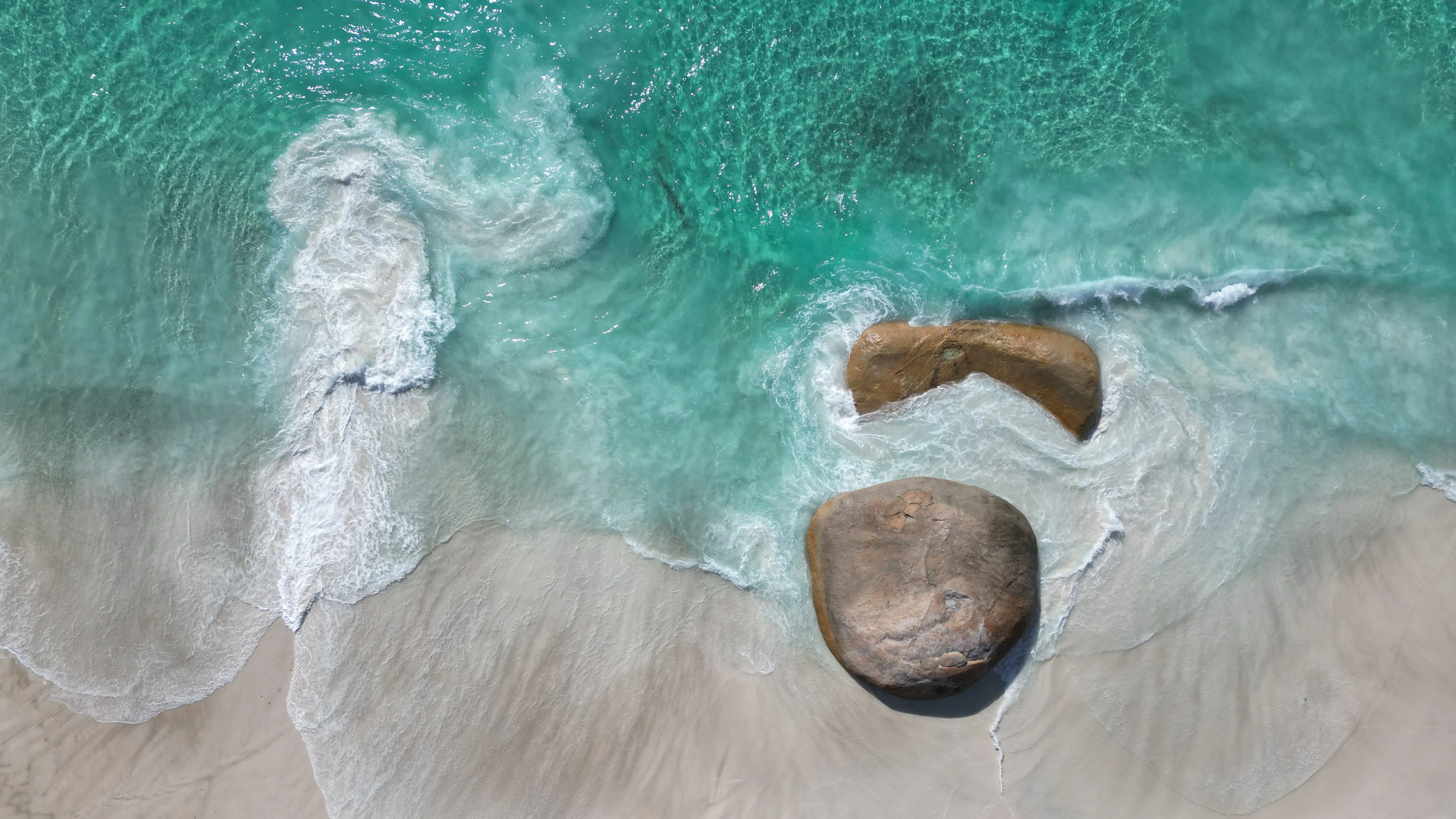 Aerial view of a beautiful beach with turquoise waters, white sand, and large rocks, showcasing a premium coastal holiday destination ideal for guests seeking a relaxing getaway, fully managed for an effortless holiday experience