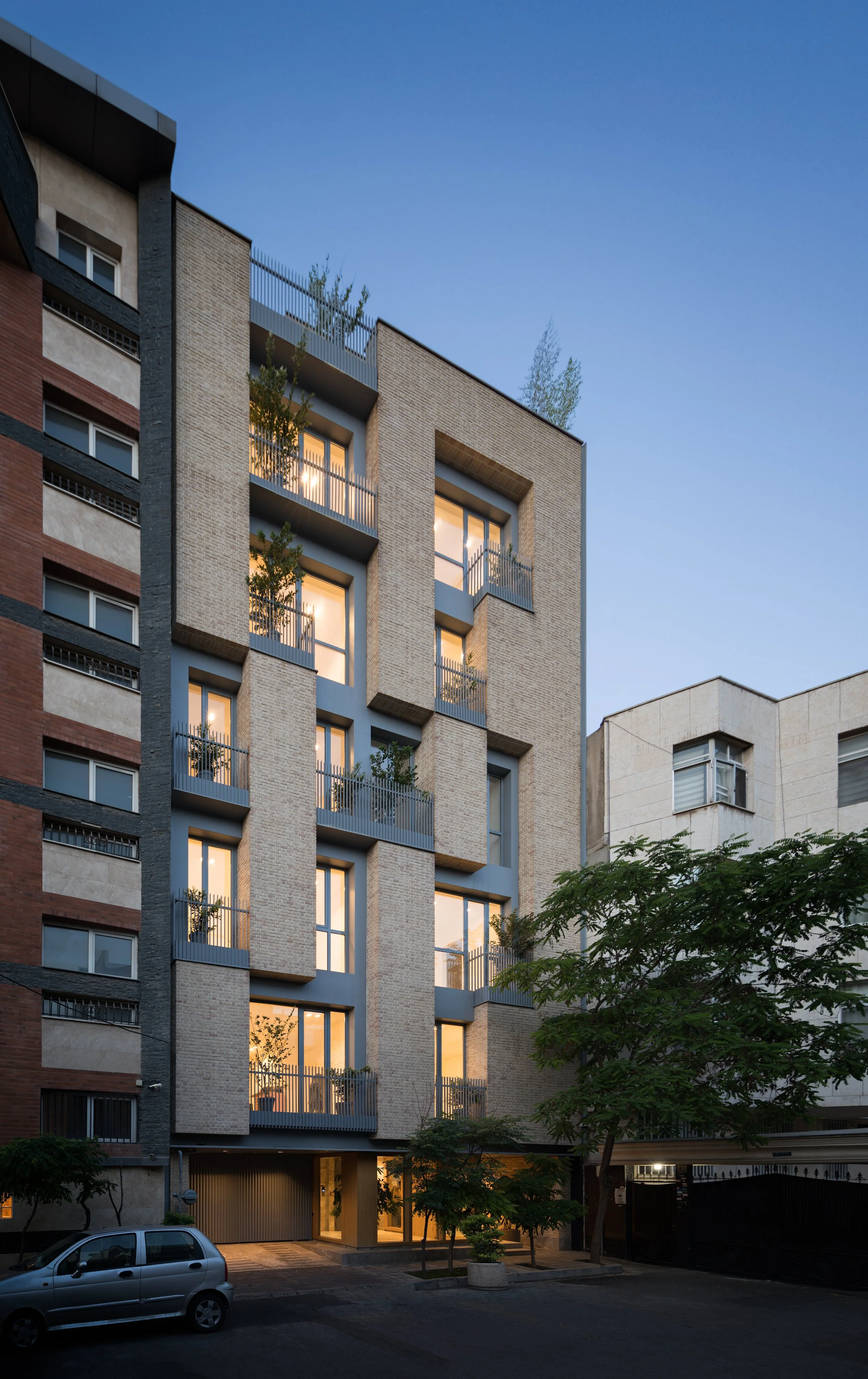 Modern multi-story apartment building with illuminated windows and balconies, trees in front, parked car, and a clear evening sky.