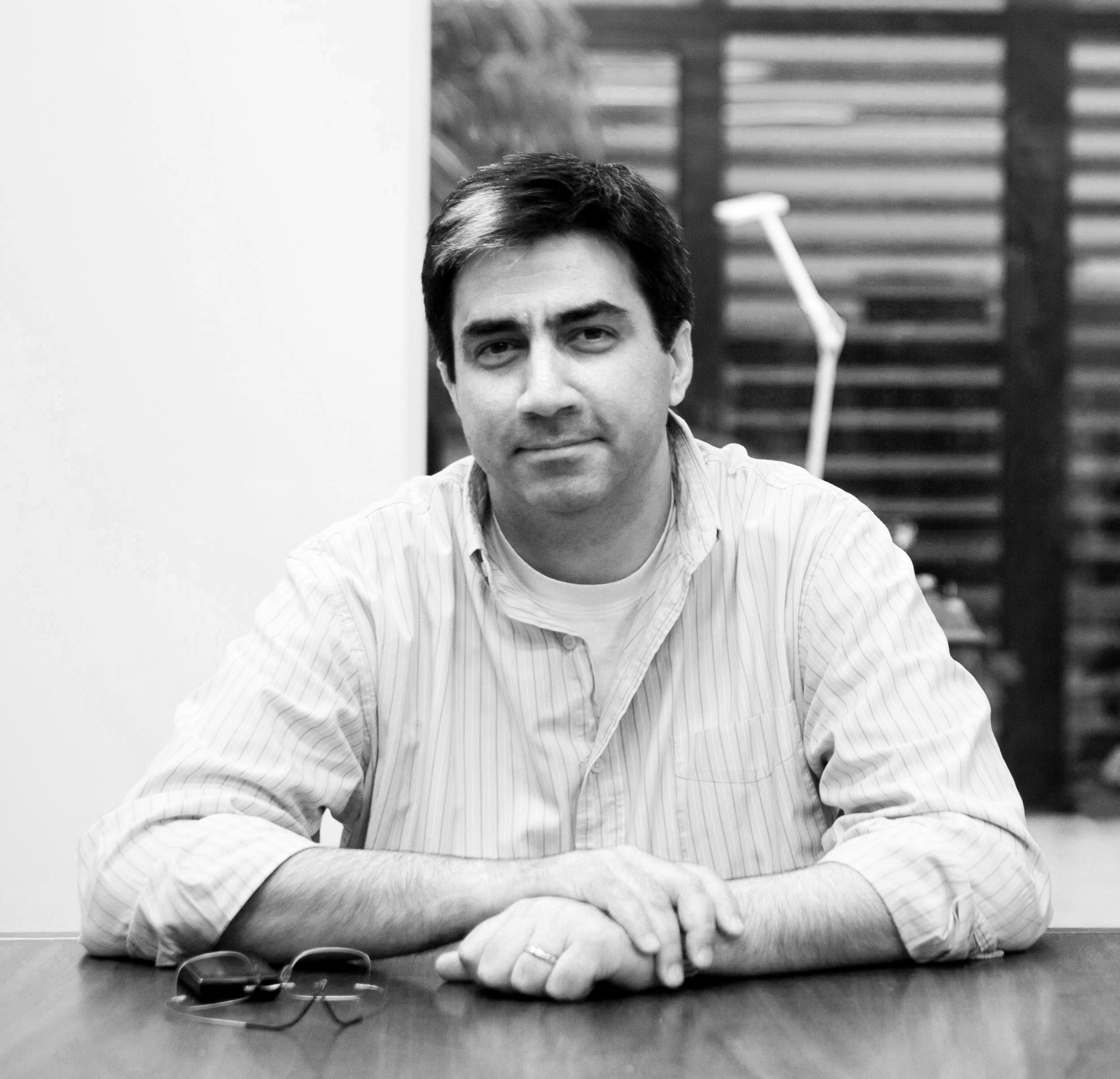 Black and white photograph of a man with dark hair and light skin, sitting at a desk with his chin resting on his crossed arms. He is wearing a striped shirt and looking directly at the camera. A pair of glasses and a wristwatch are on the desk. The background shows wooden slats and a white wall.