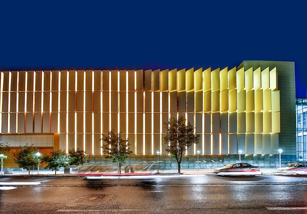 Nighttime view of a modern building with illuminated yellow and white slats, trees, parking lot, and moving cars.