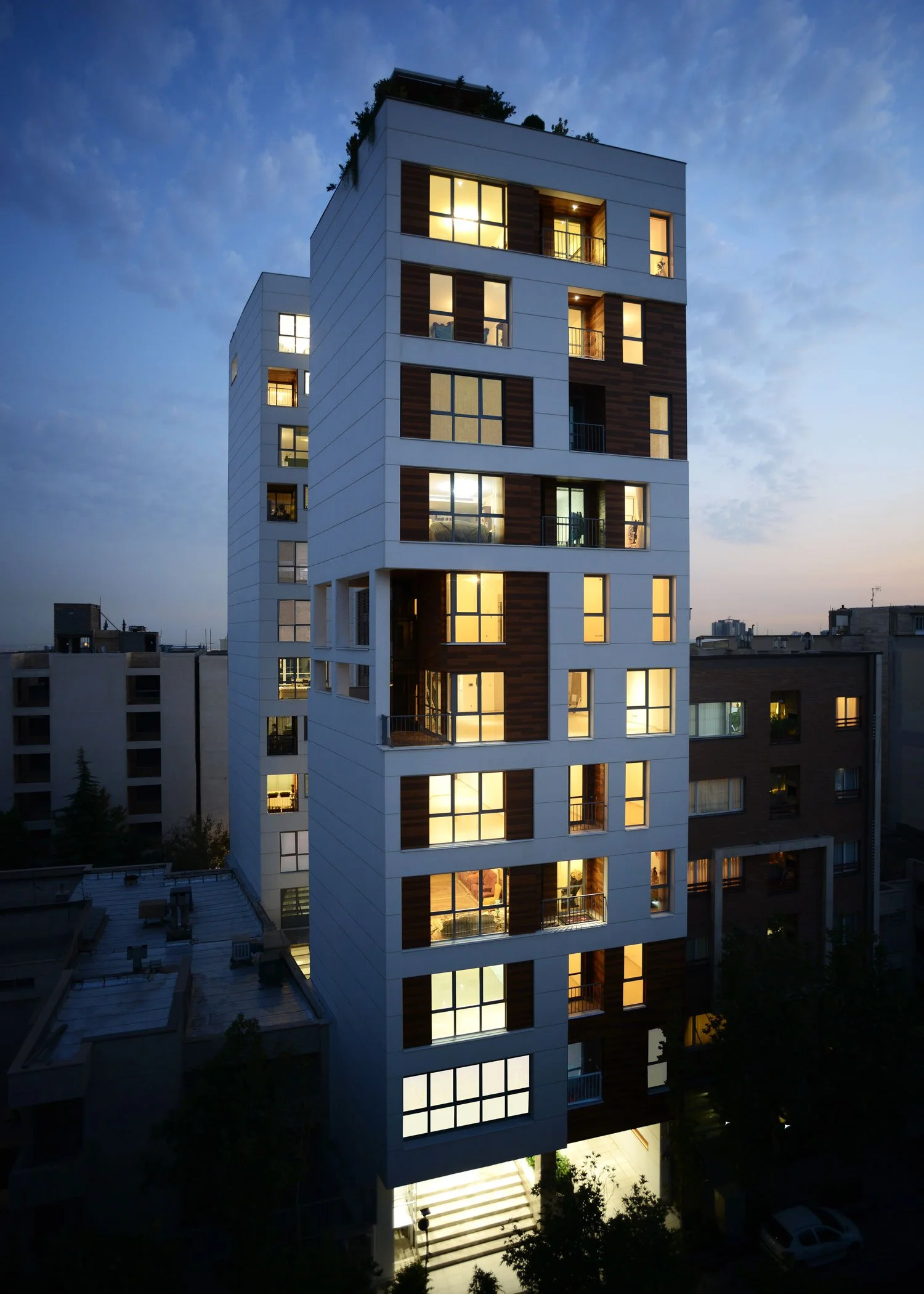 A modern multi-story residential building at dusk with illuminated windows and a staircase leading to the entrance.
