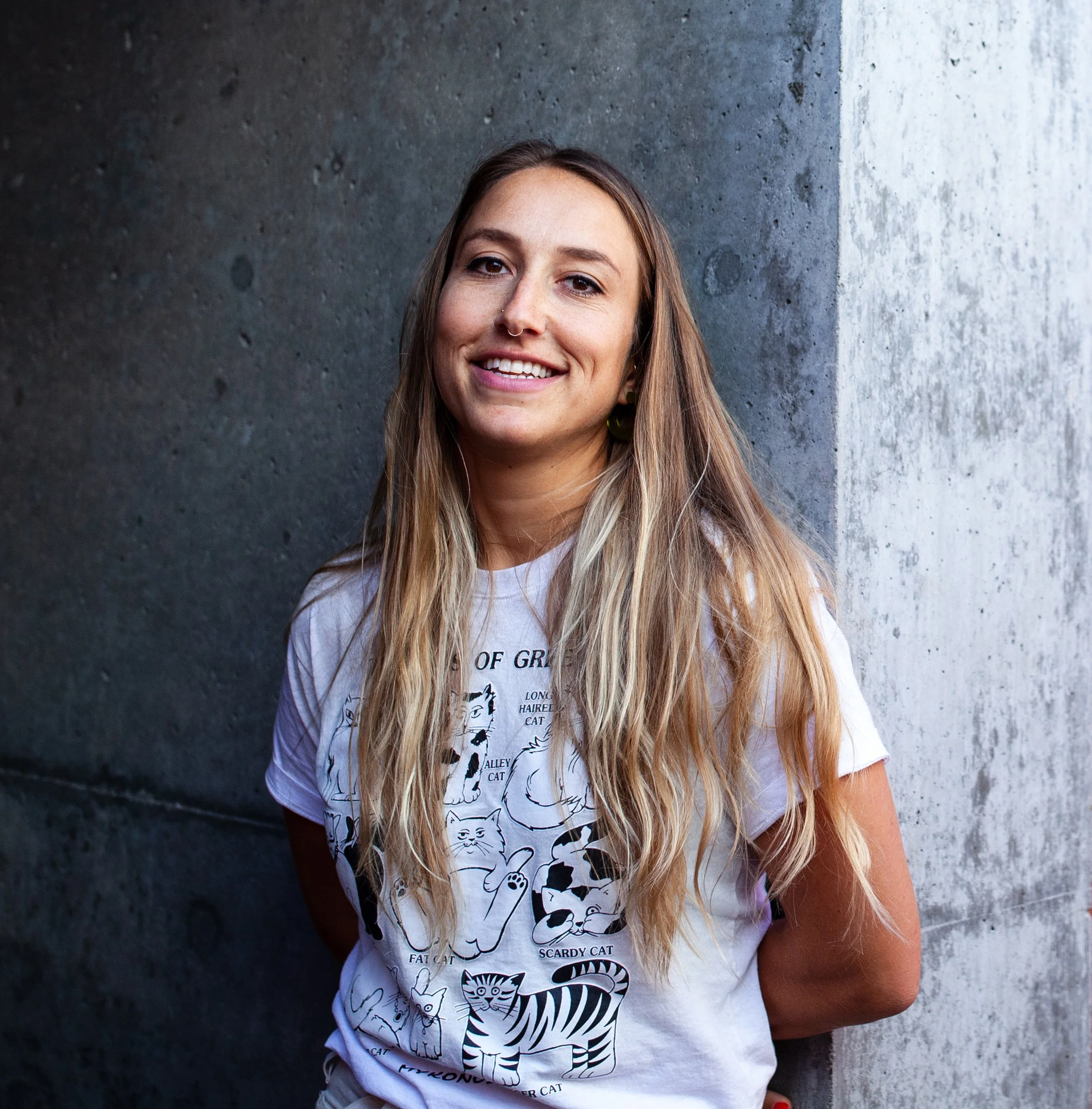 Portrait of the artist Alice Wiese in front of a textured gray wall wearing a cat t-shirt for Soapbox Arts Gallery in Burlington, Vermont