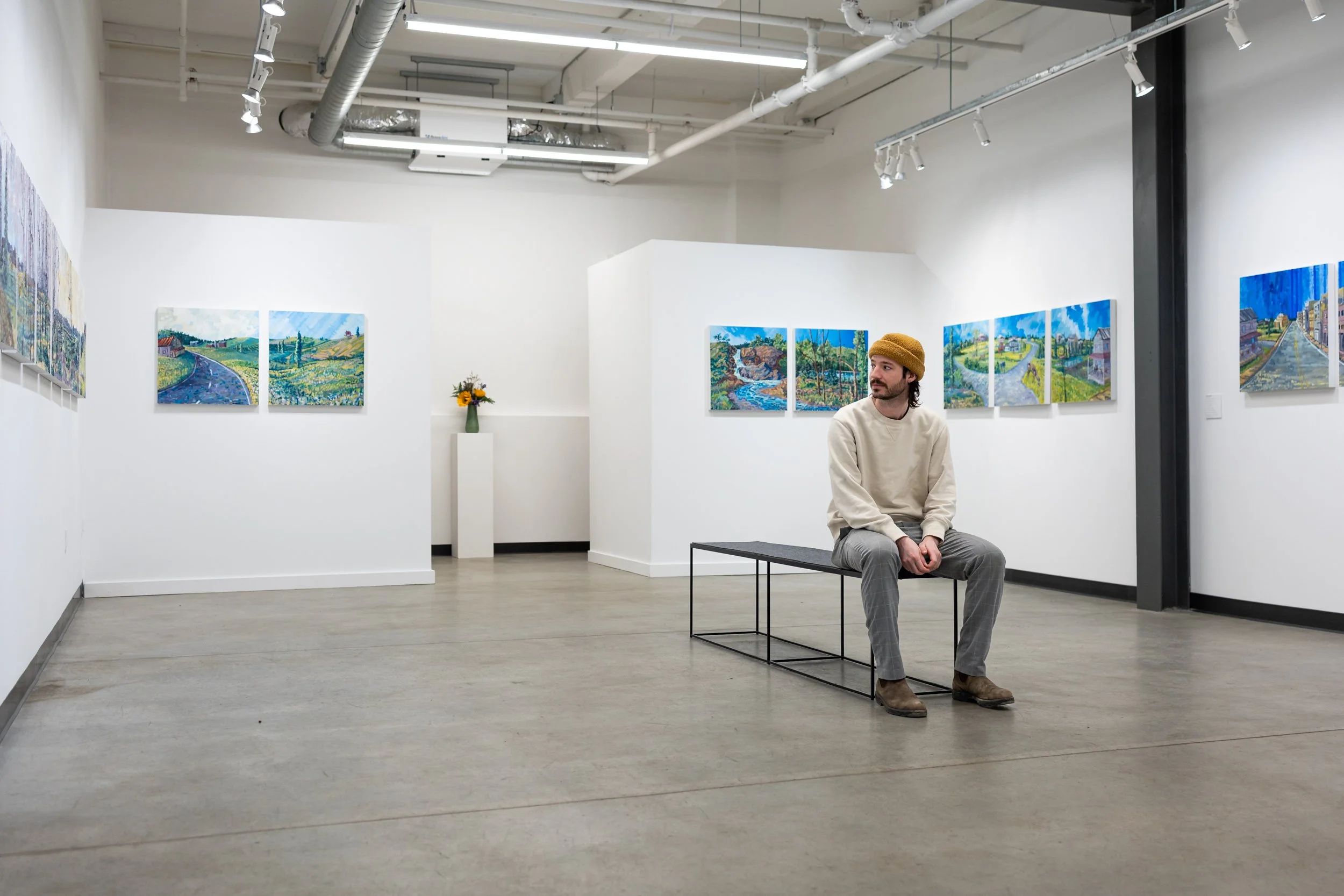 the artist Charlie Hudson sitting in the gallery surrounded by an installation of his landscape paintings at Soapbox Arts Gallery in Burlington, Vermont