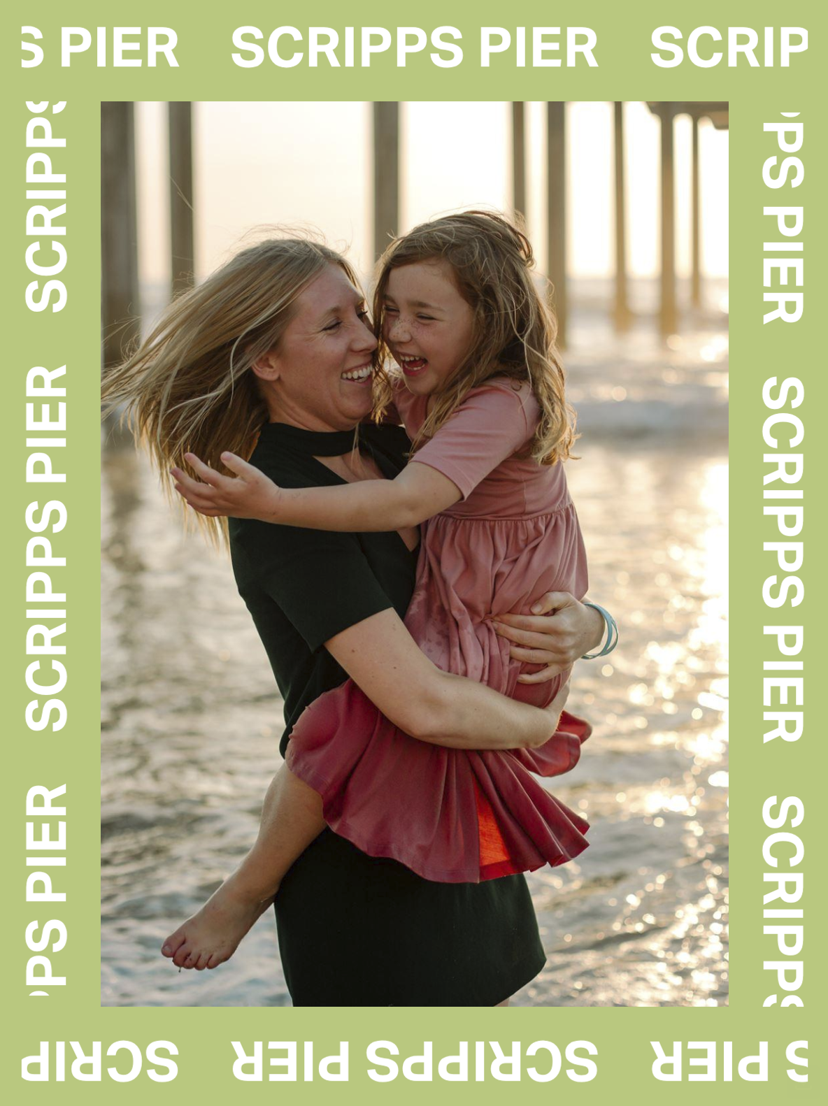 A woman holding a young girl in her arms, both smiling and laughing near a body of water with a pier in the background during sunset.