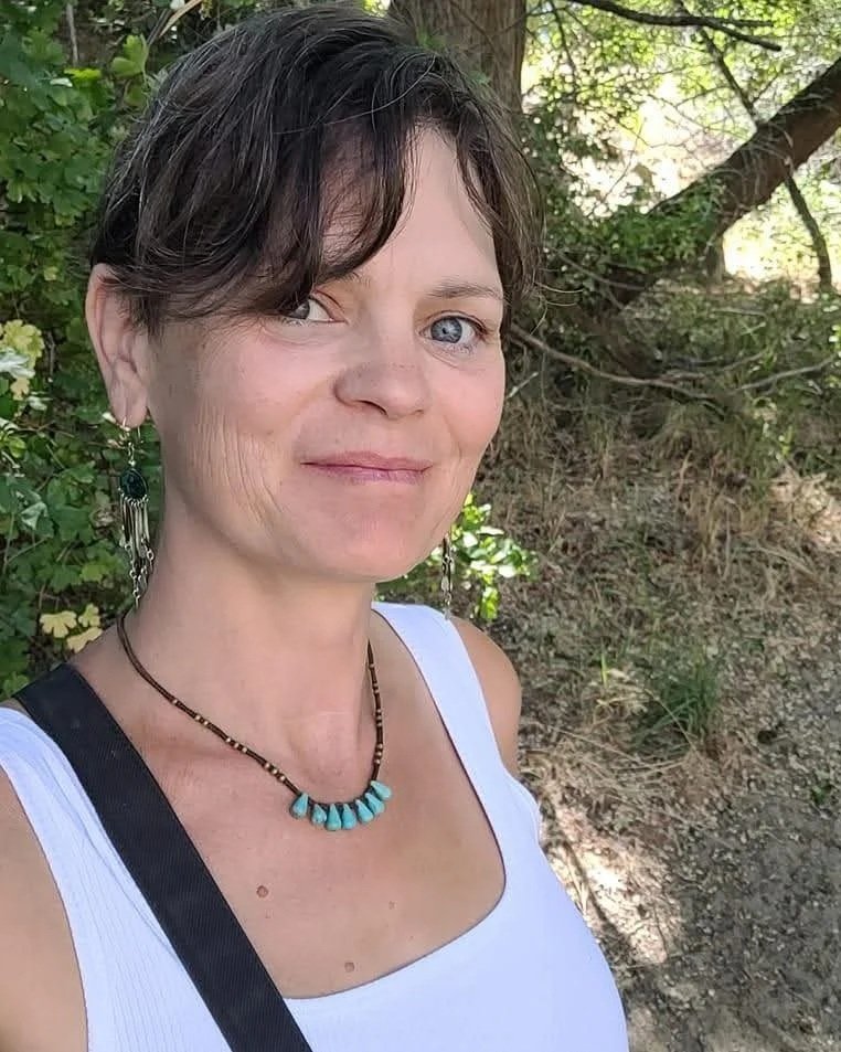 woman sam switzer looking at camera wearing white vest standing in a wood