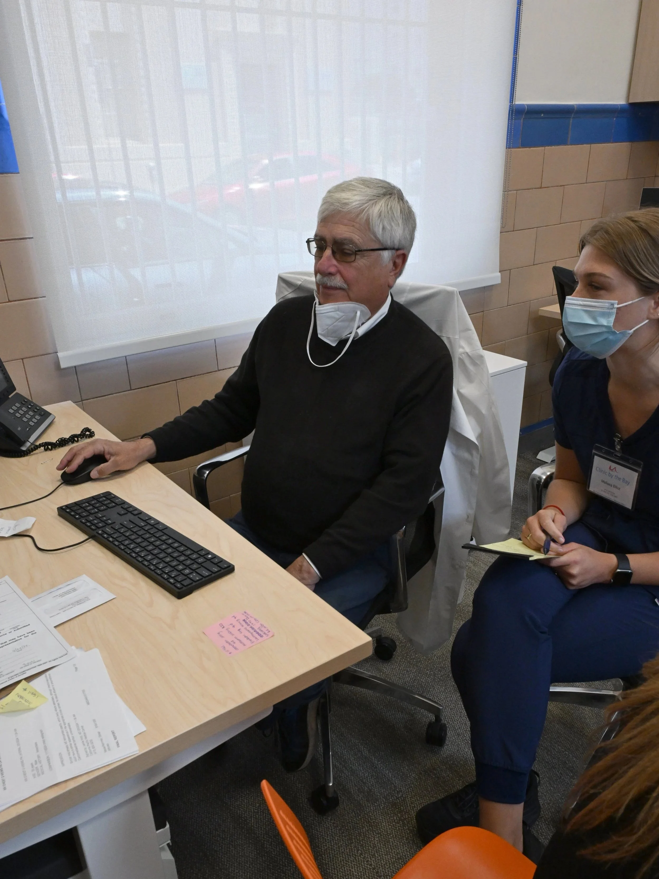 Two women sitting at a desk in an office, smiling. The desk has computer monitors, papers, masks, and other office supplies. Behind them is a blue tiled wall with framed documents, a globe, and stacks of folders. A photocopy machine is also visible.
