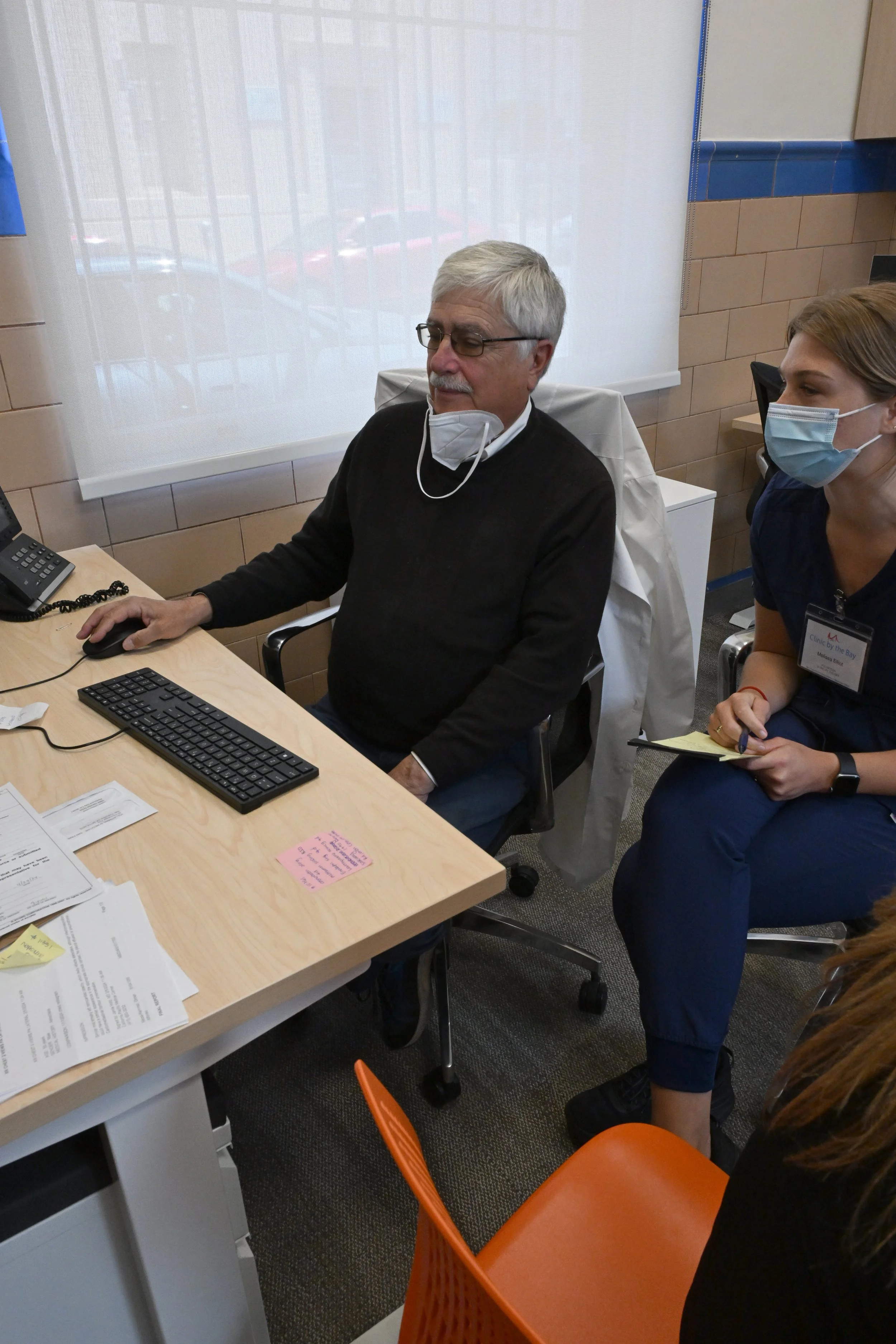 Two women sitting at a desk in an office, smiling. The desk has computer monitors, papers, masks, and other office supplies. Behind them is a blue tiled wall with framed documents, a globe, and stacks of folders. A photocopy machine is also visible.