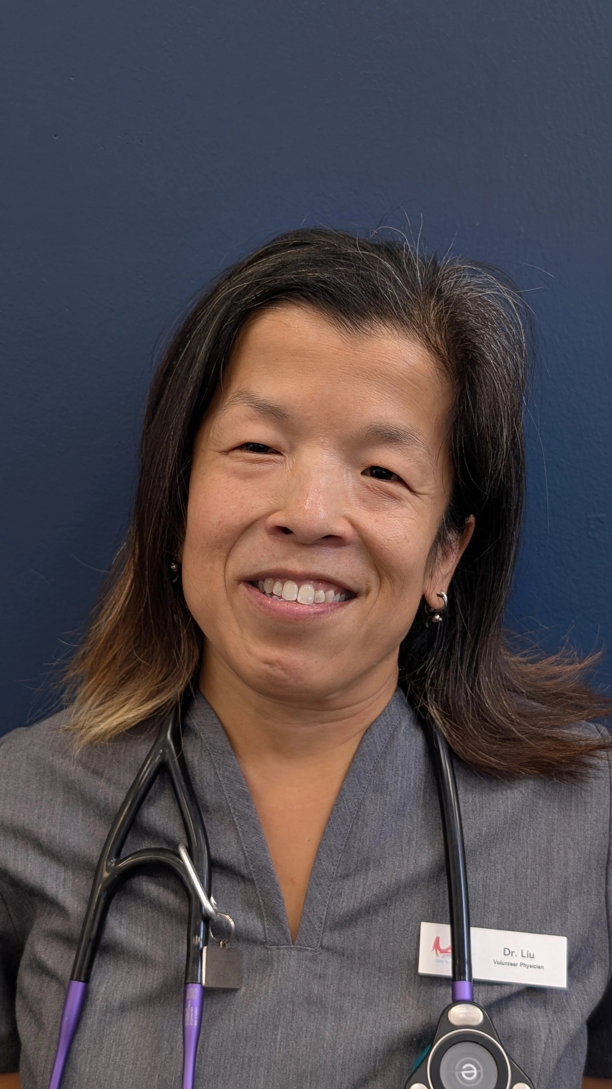 A smiling woman with long dark hair, wearing gray medical scrubs, a stethoscope, and a name tag, standing against a dark blue wall.