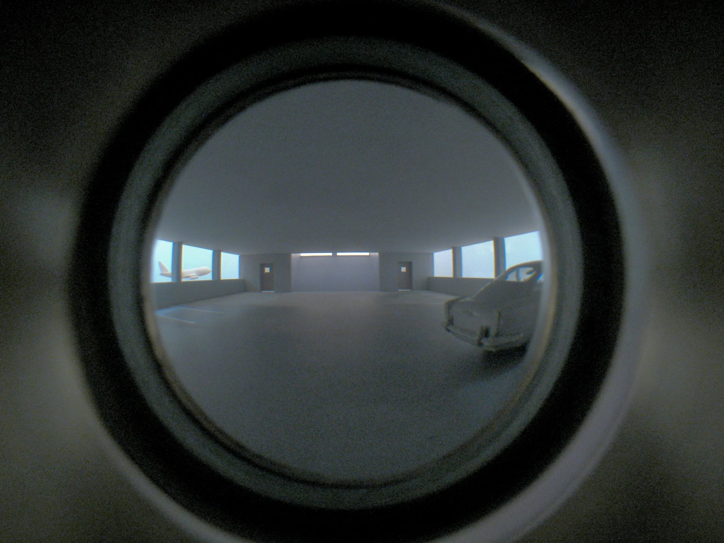 Airport Parking Garage , interior view Looking through a peephole lens, the viewer glimpses an empty parking garage. Approximately every 25 seconds, an airplane slowly glides past. A recording, composed of looped found sound, is loosely synced to the motion of the plane. Materials include: MDF, acrylic, polystyrene, Plexiglas, halogen lights, 12 RPM gear motor, belt, paperclip, sound component (looped recording