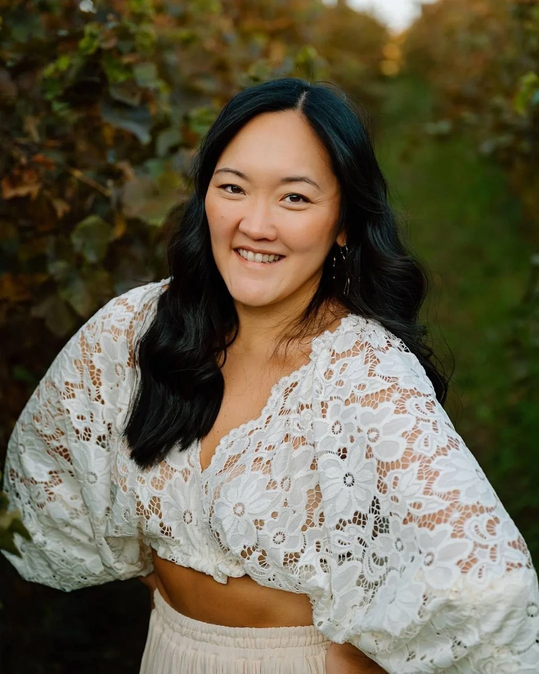 Women with dark hair wearing a white lace top and white skirt in a vineyard.