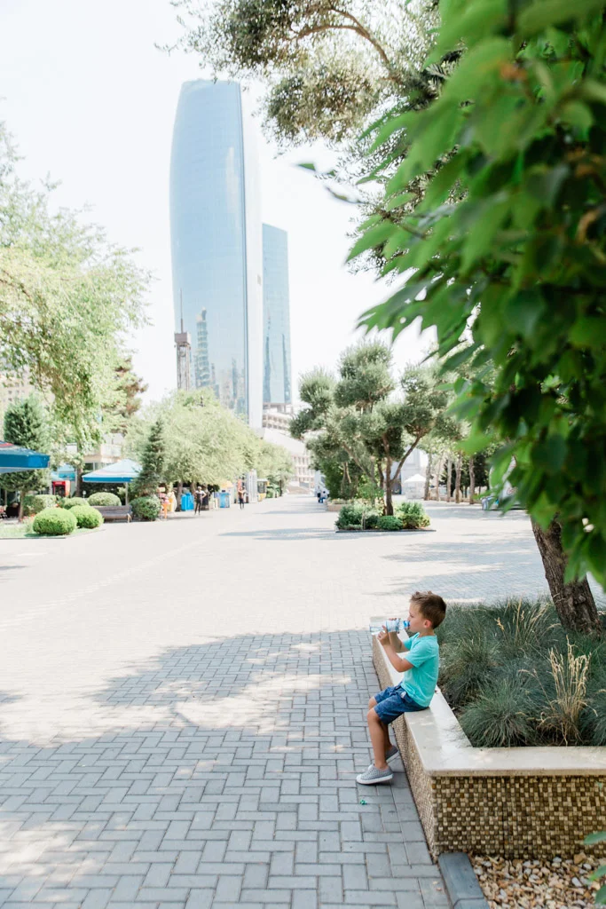 Little boy drinking from a water bottle sitting on the edge of a planter in a park with skyscraper in the background.