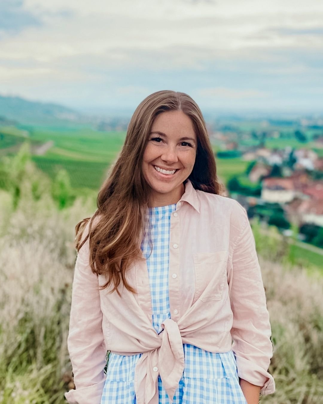 Woman with a blue and white gingham dress and pink button down shirt with German village in background.