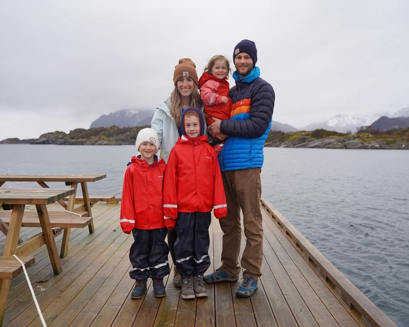 Family of 5 in cold weather clothes on a dock with cloudy mountains in the backround