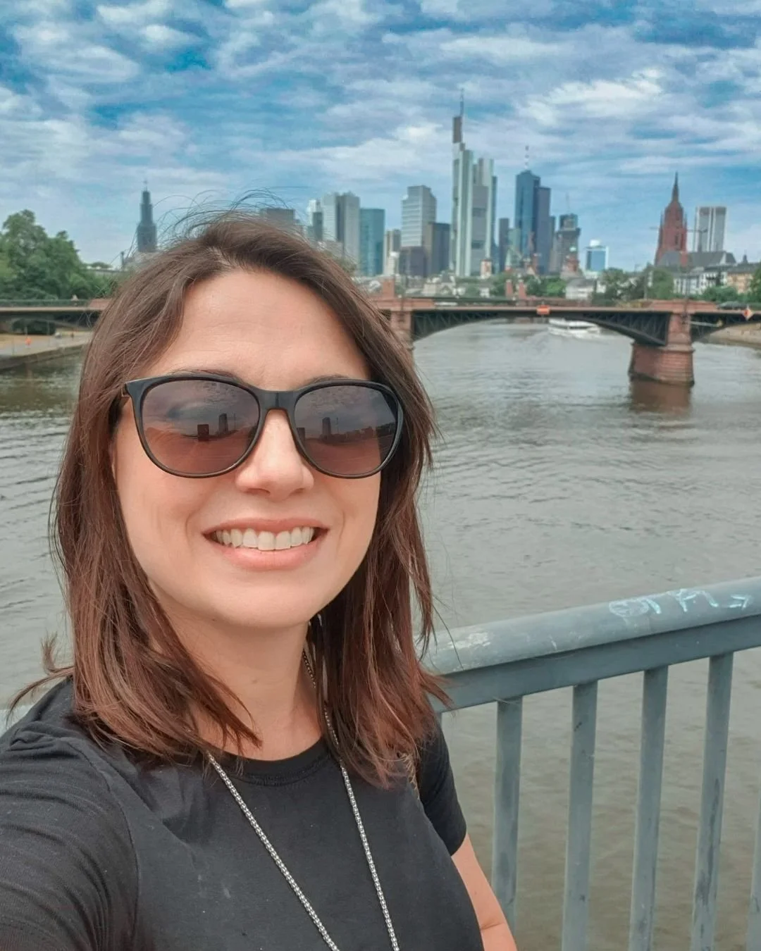 Selfie of woman in black t-shirt and sunglasses on a bridge over a river in Frankfurt, Germany