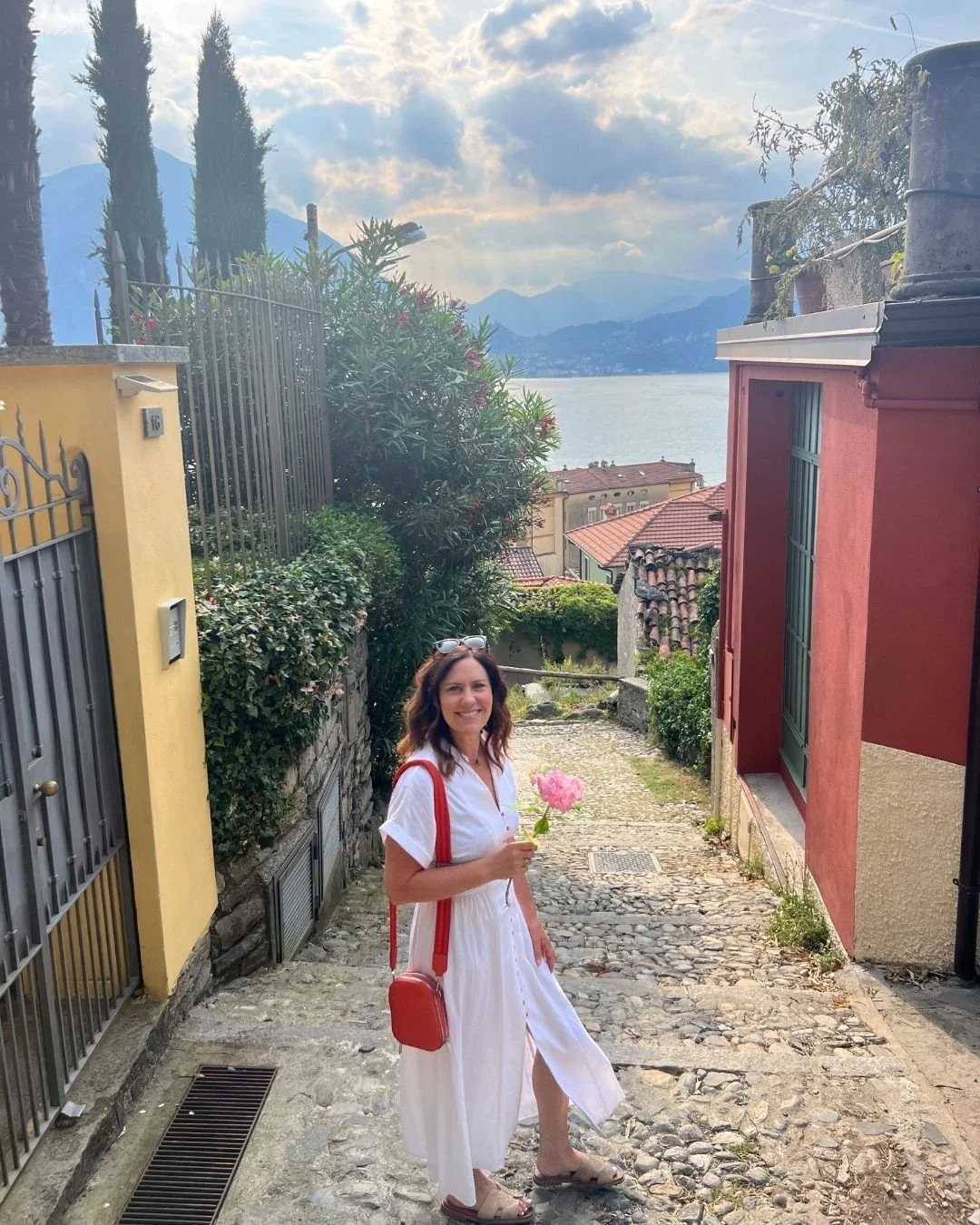 Woman in white dress holding a pink flower in a small cobblestone alley with lake and mountains in the background.