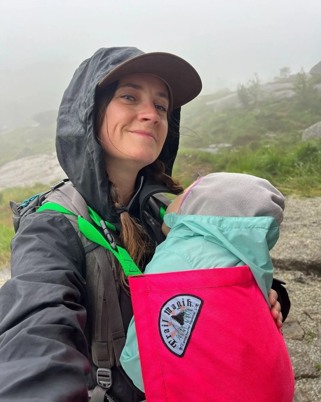 Selfie of a woman hiking wearing a baby in a pink baby carrier both wearing rain coats