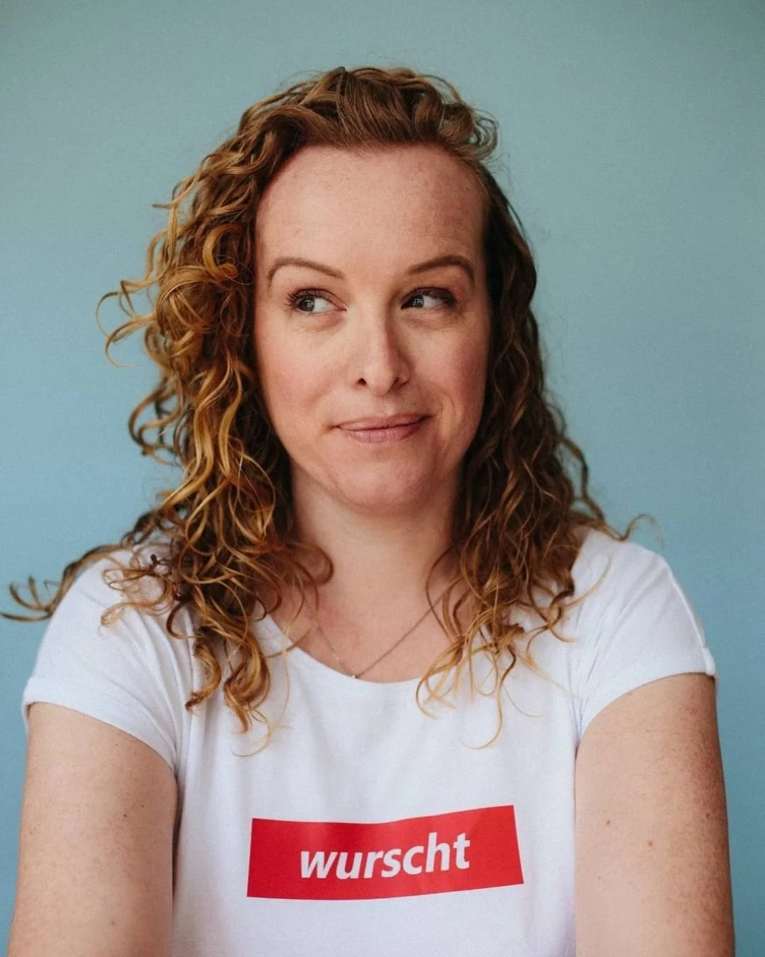 Light teal backdrop with a close up of a woman with red, curly hair looking to the left. Her white t-shirt says, "wurscht"