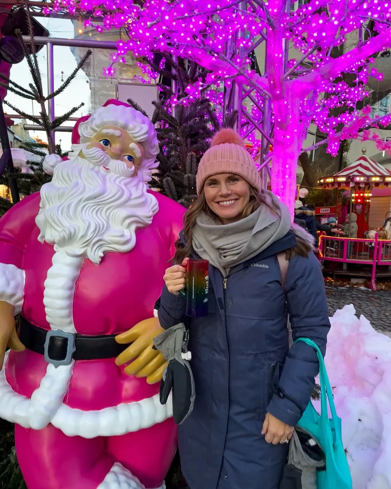 Woman in a blue winter coate and pink hat with a rainbow Christmas market mug next to a pink Santa in Munich, Germany