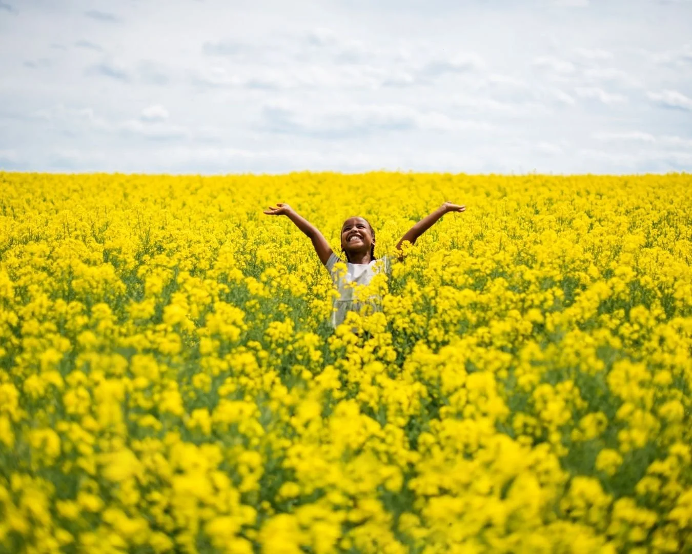 Little girl in a field of yellow flowers reaching up with arms outstretched and smiling.