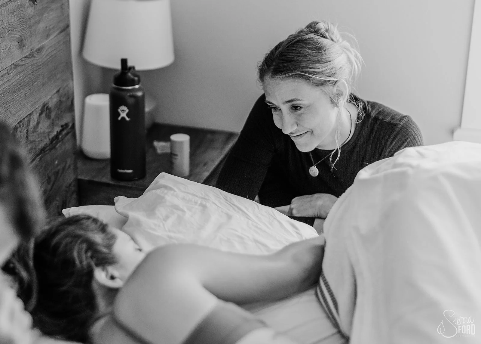 A woman with blonde hair up in a bun sitting beside a bed, smiling and looking at a child lying in bed with their arm resting on the blanket. The child's face is turned away from the camera.